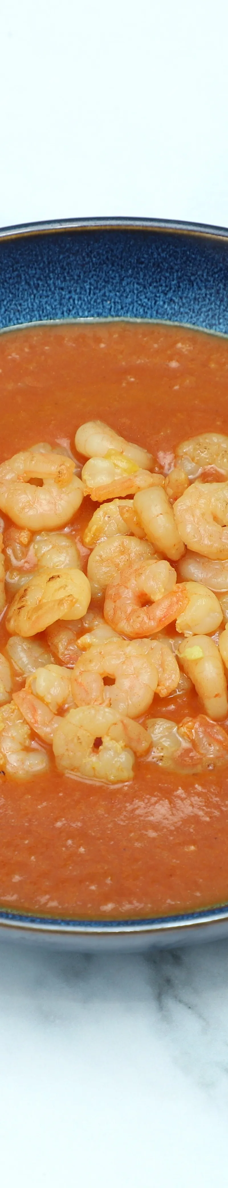 Top-down view of a dark blue bowl filled with orange-red curry and many small cooked shrimp.