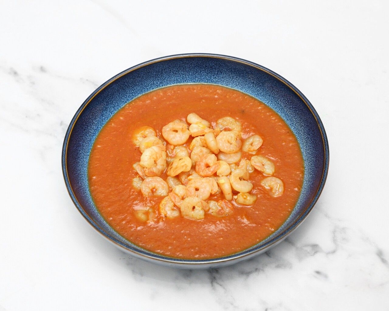Small peeled prawns in a vibrant orange-red curry sauce, served in a dark blue bowl.
