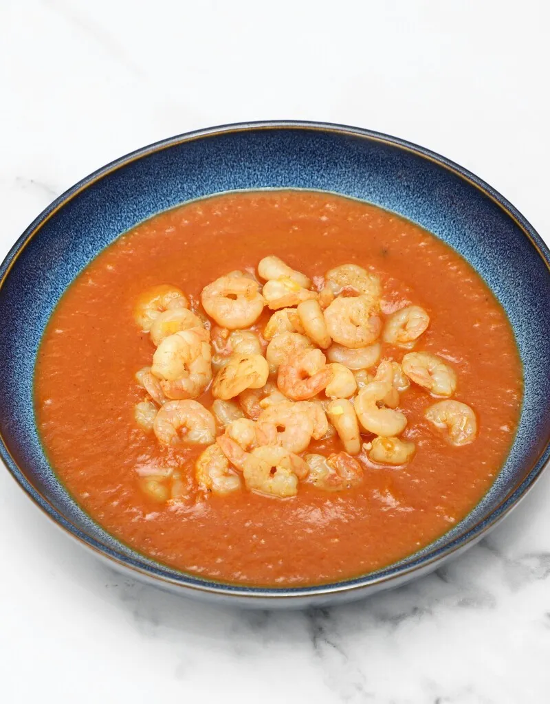 Small peeled prawns in a vibrant orange-red curry sauce, served in a dark blue bowl.