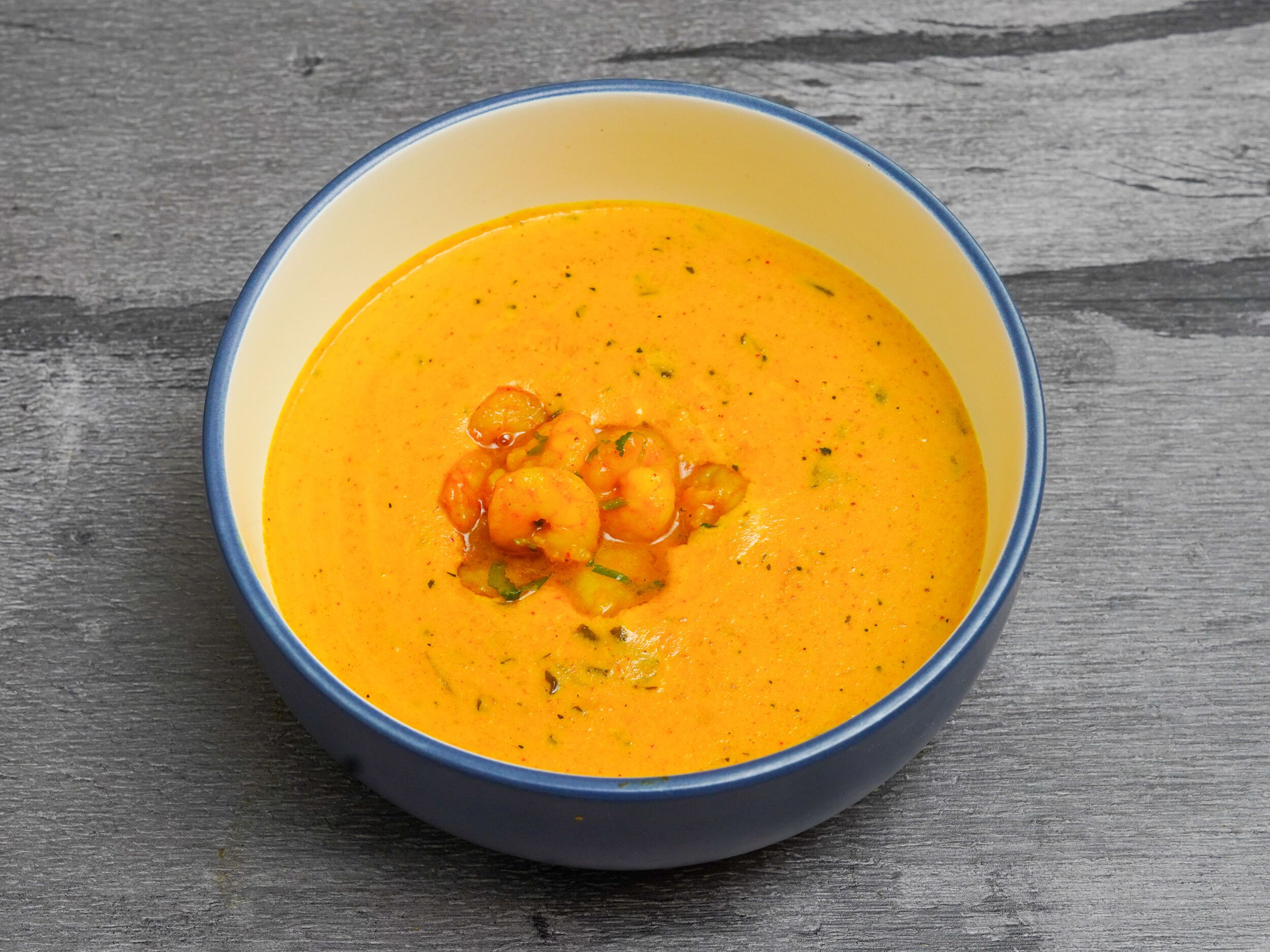 Orange prawn curry with visible herbs and spices in a blue and cream bowl on a grey wooden surface.