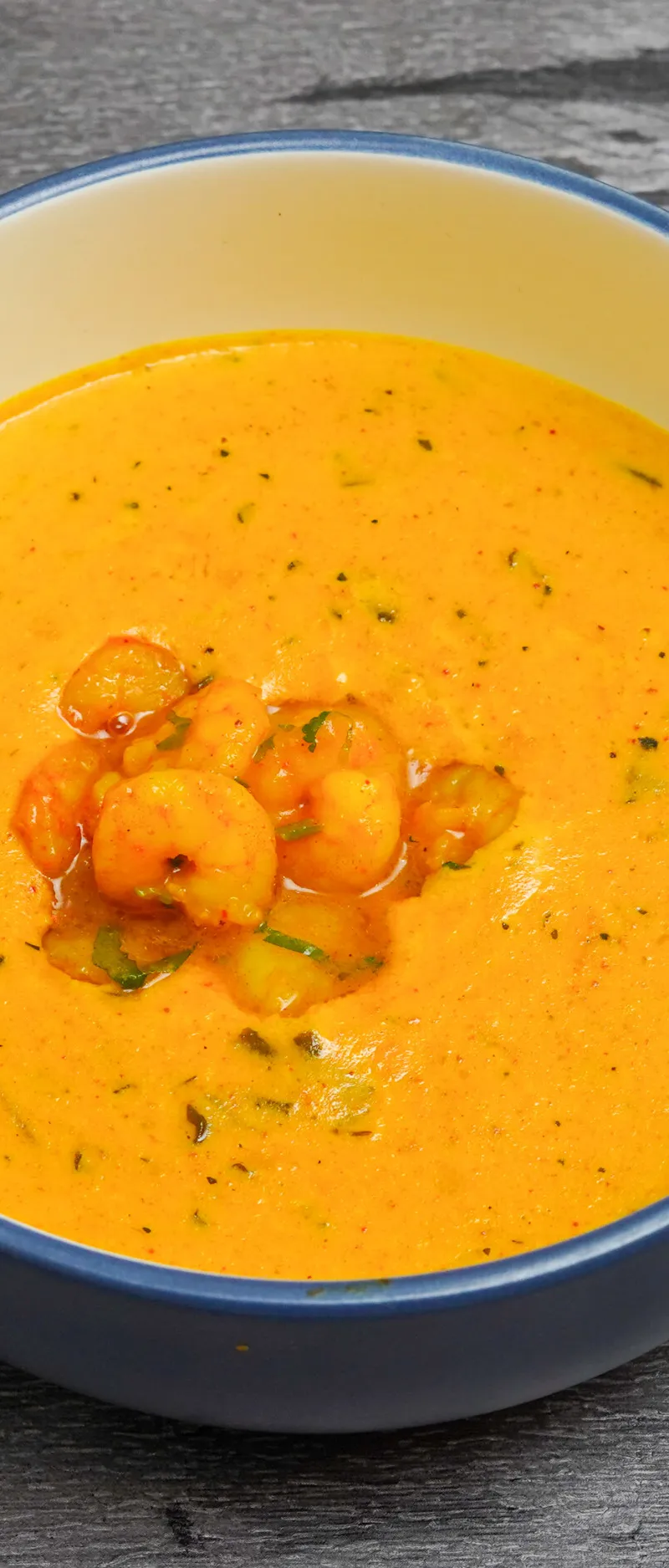 Orange prawn curry with visible herbs and spices in a blue and cream bowl on a grey wooden surface.