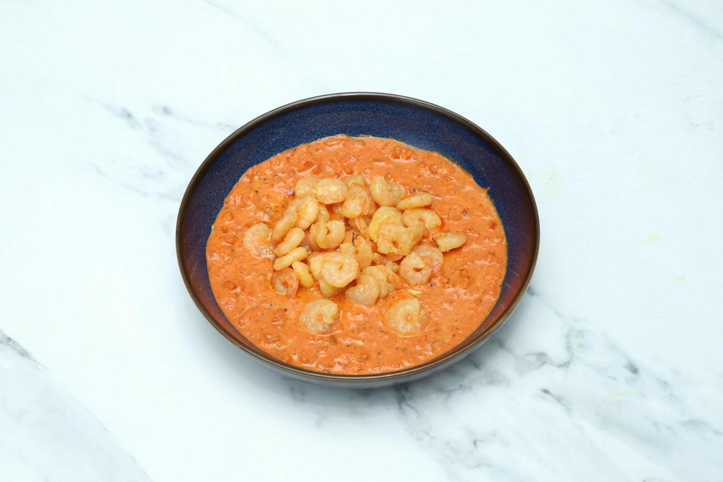 A bowl of king prawn butterfly in a rich, orange-red curry sauce on a white marble background.