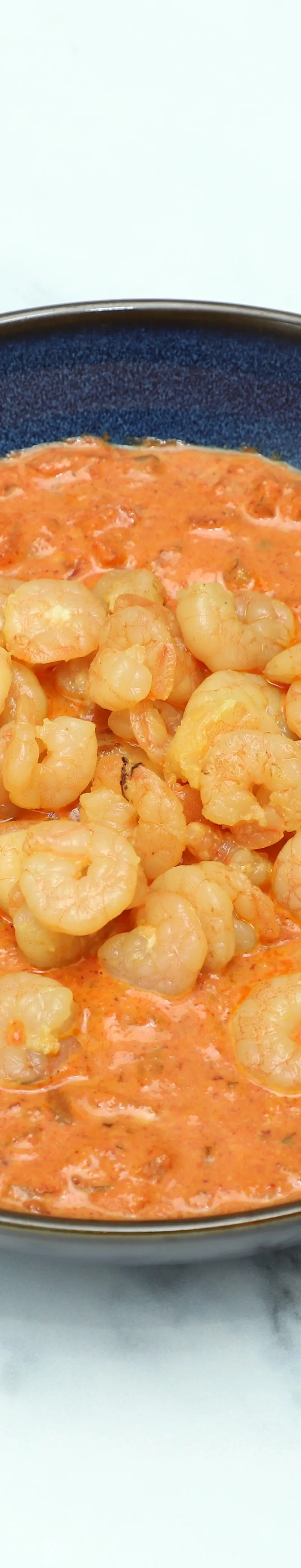 A bowl of king prawn butterfly in a rich, orange-red curry sauce on a white marble background.