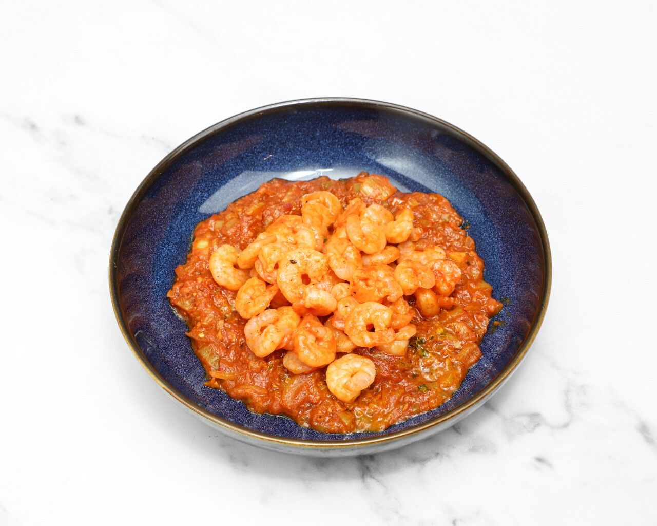 A deep blue bowl filled with small cooked shrimp in a rich, reddish-orange curry sauce,
