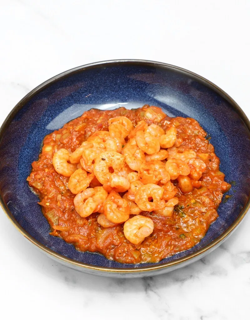 A deep blue bowl filled with small cooked shrimp in a rich, reddish-orange curry sauce,