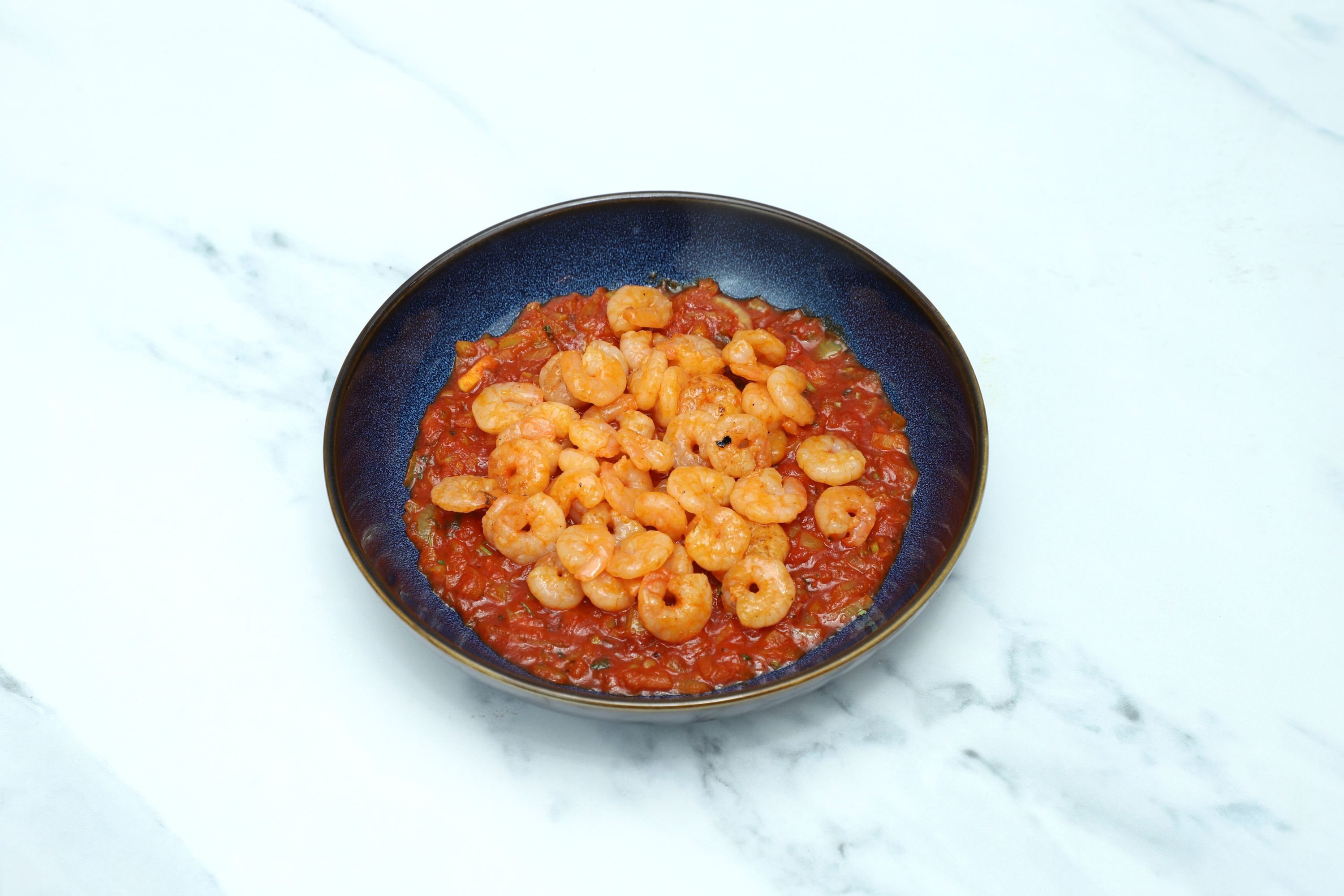 Small cooked shrimp are arranged on a deep blue bowl of red tomato curry sauce.