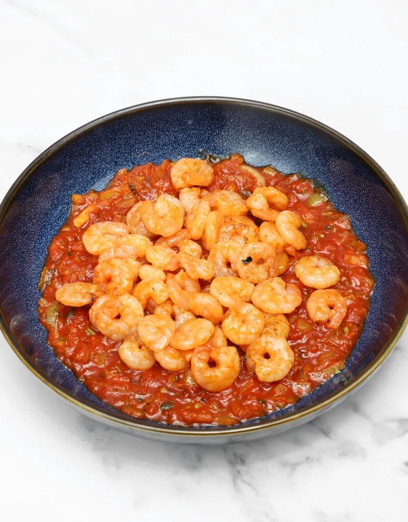 A dark blue bowl filled with small, cooked shrimp in a vibrant red tomato sauce.