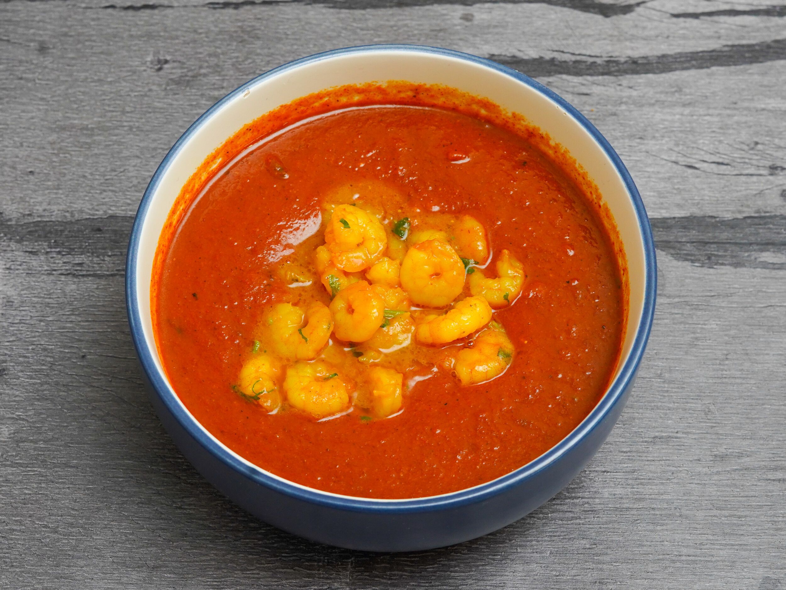 A blue and white bowl filled with a rich orange prawn curry, against a grey wooden background.