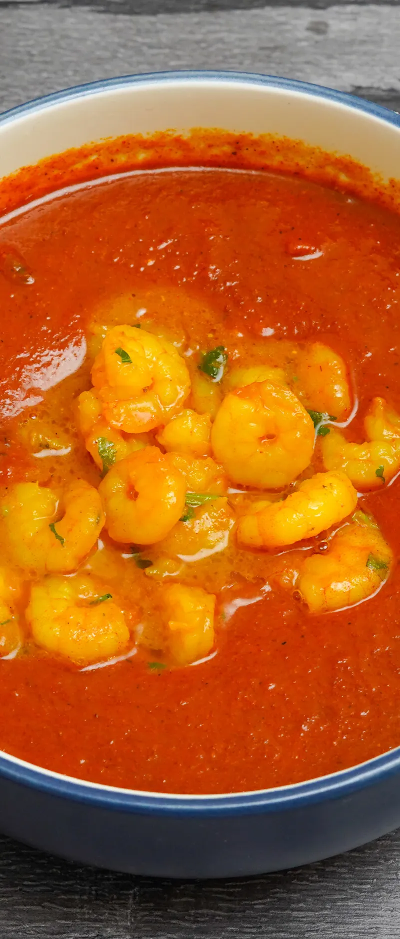 A blue and white bowl filled with a rich orange prawn curry, against a grey wooden background.