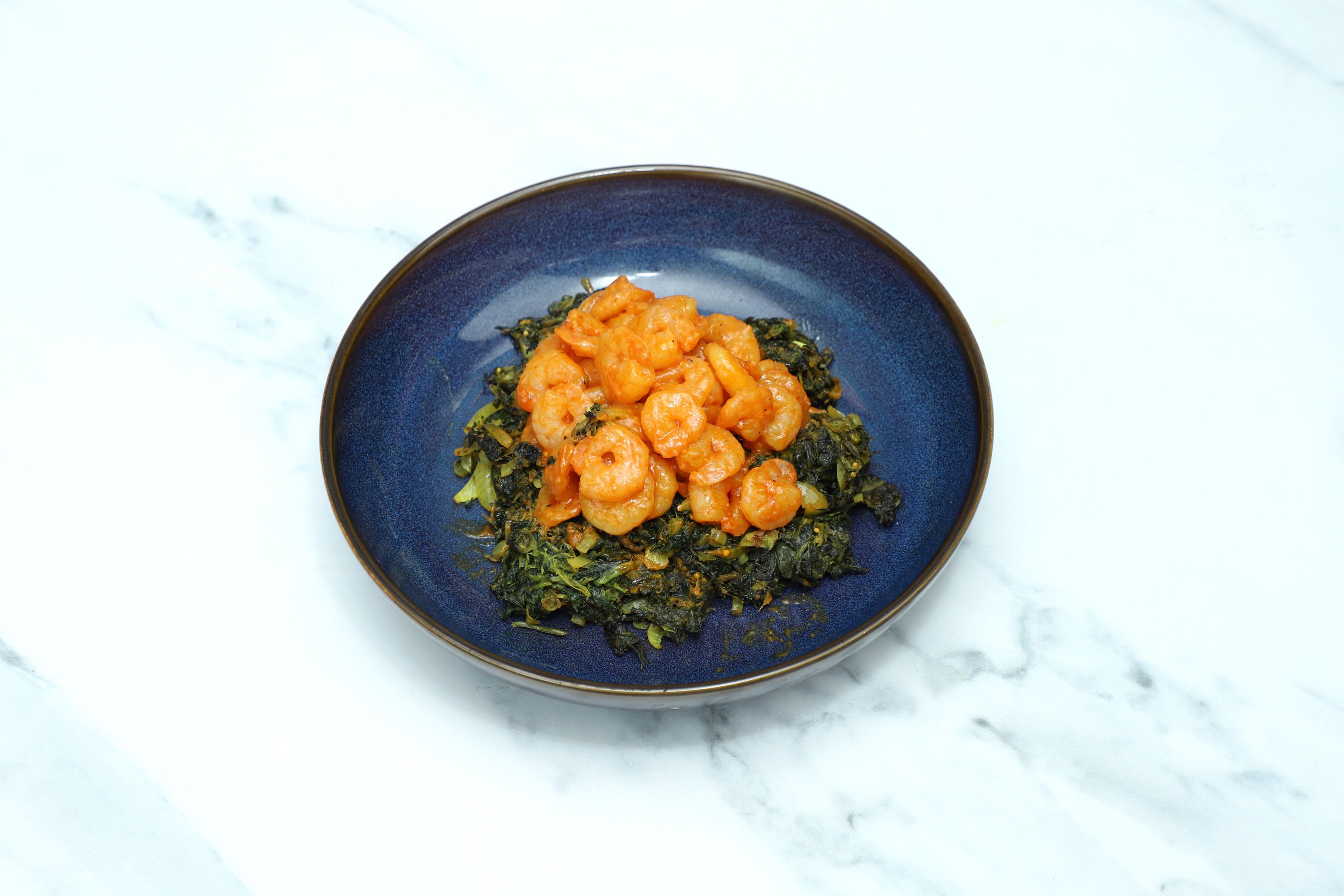 Orange shrimp curry served on top of a bed of dark green saag curry in a blue bowl