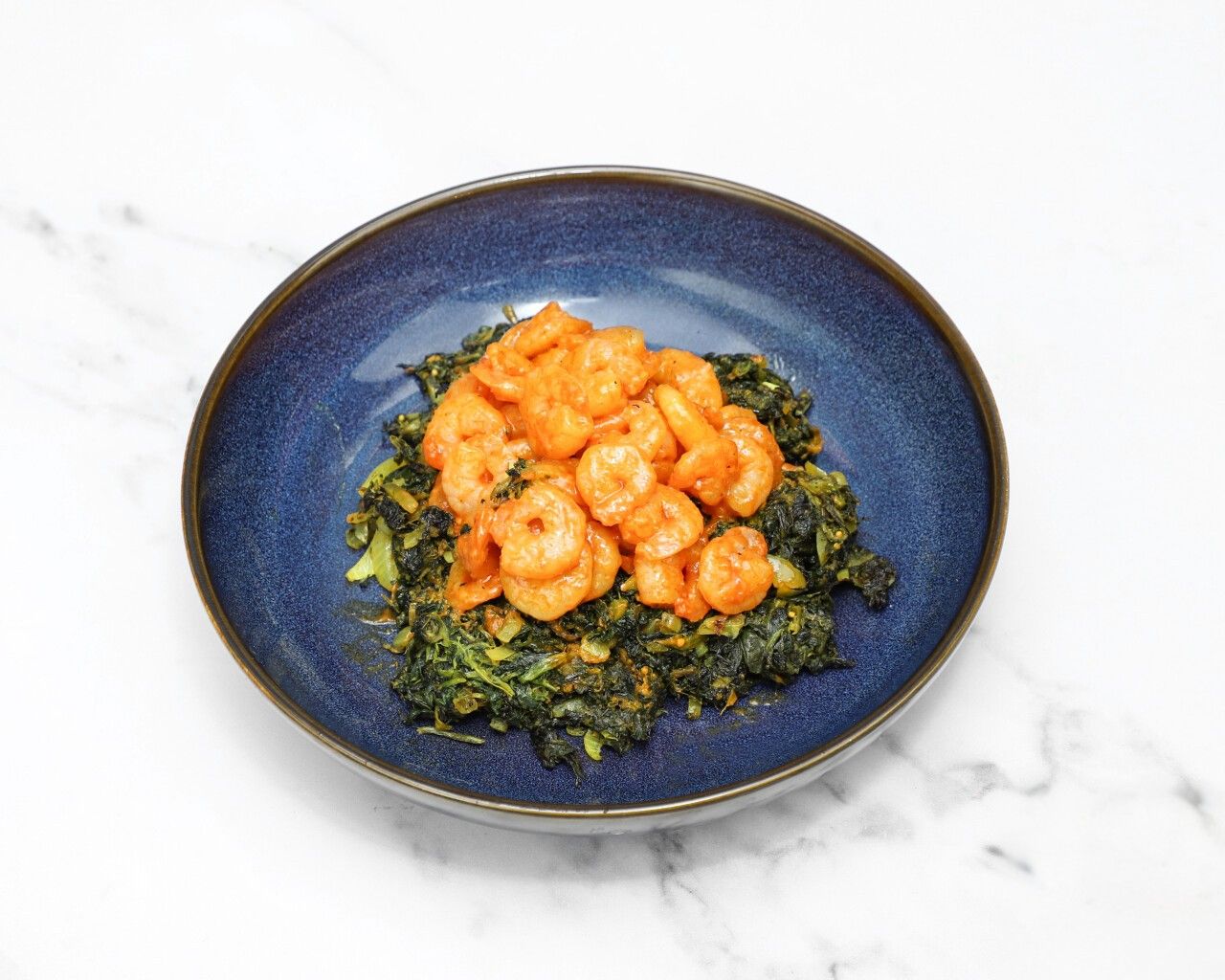 Orange prawns in sauce served atop a bed of dark green cooked spinach in a blue bowl.