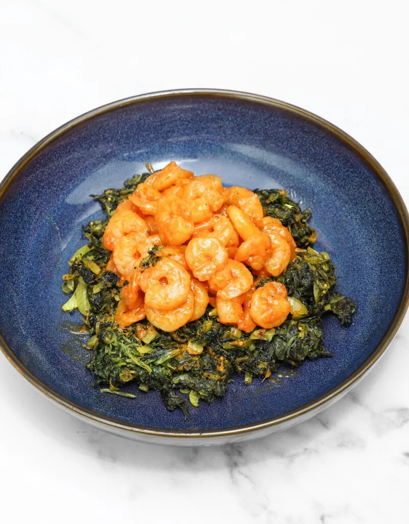 Orange prawns in sauce served atop a bed of dark green cooked spinach in a blue bowl.