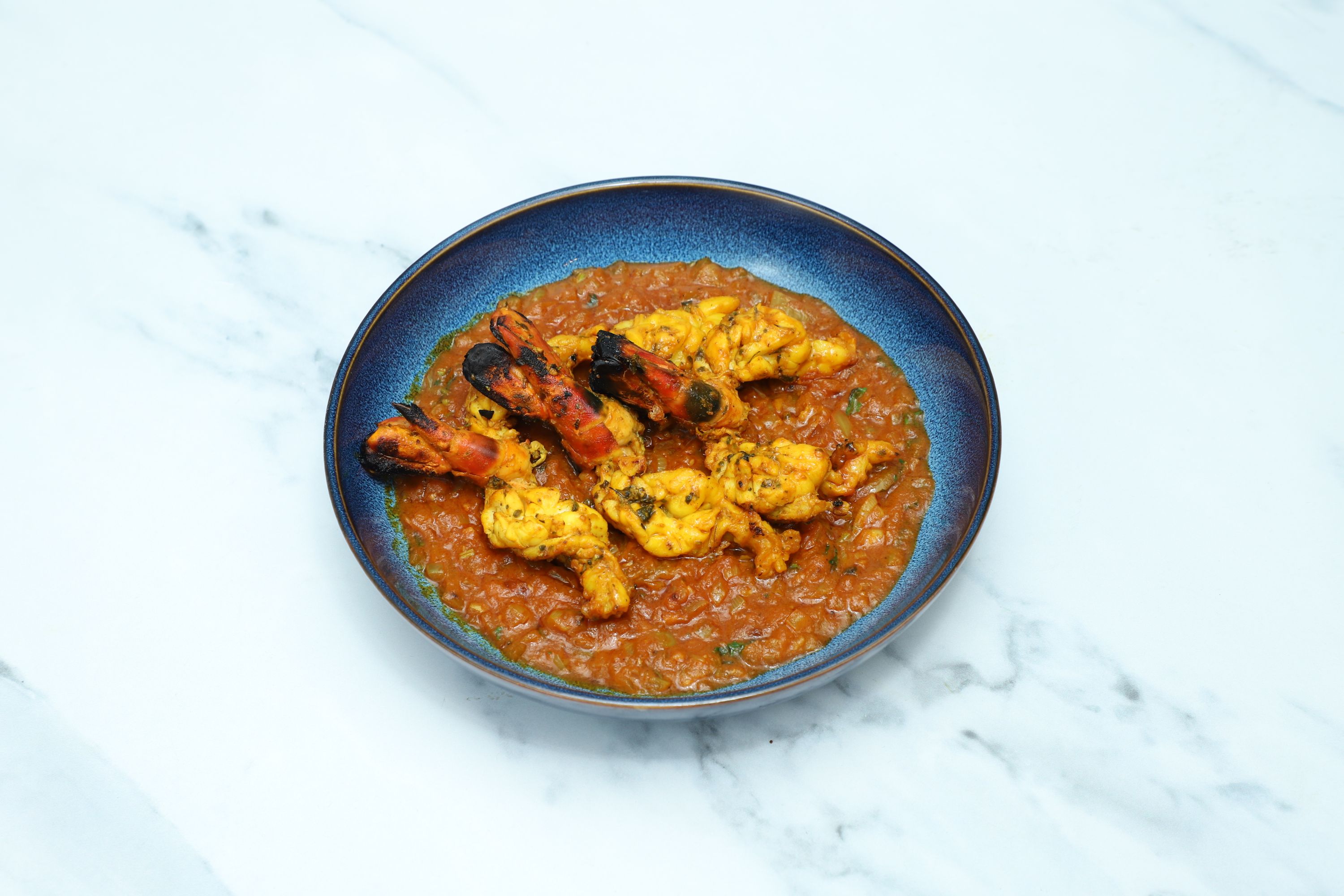 Five tandoori king prawns with charred shells arranged on a generous bed of orange curry sauce in a blue bowl.