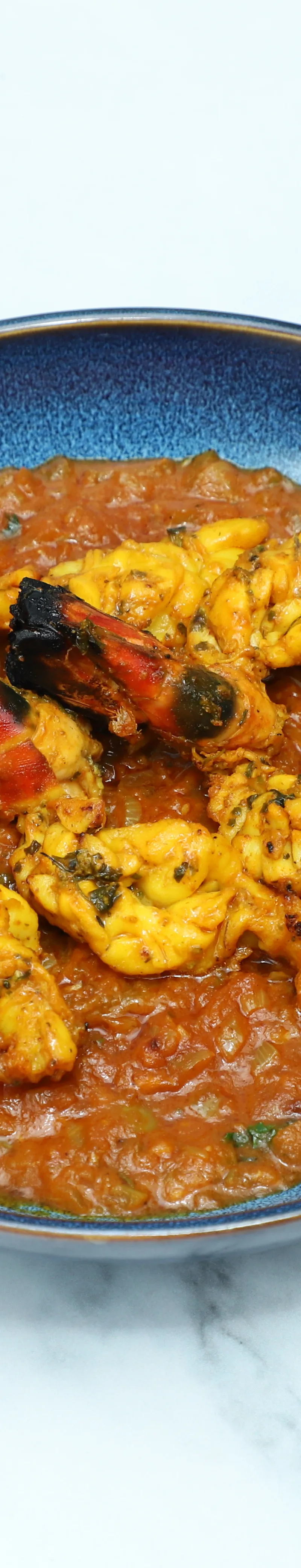 Five tandoori king prawns with charred shells arranged on a generous bed of orange curry sauce in a blue bowl.