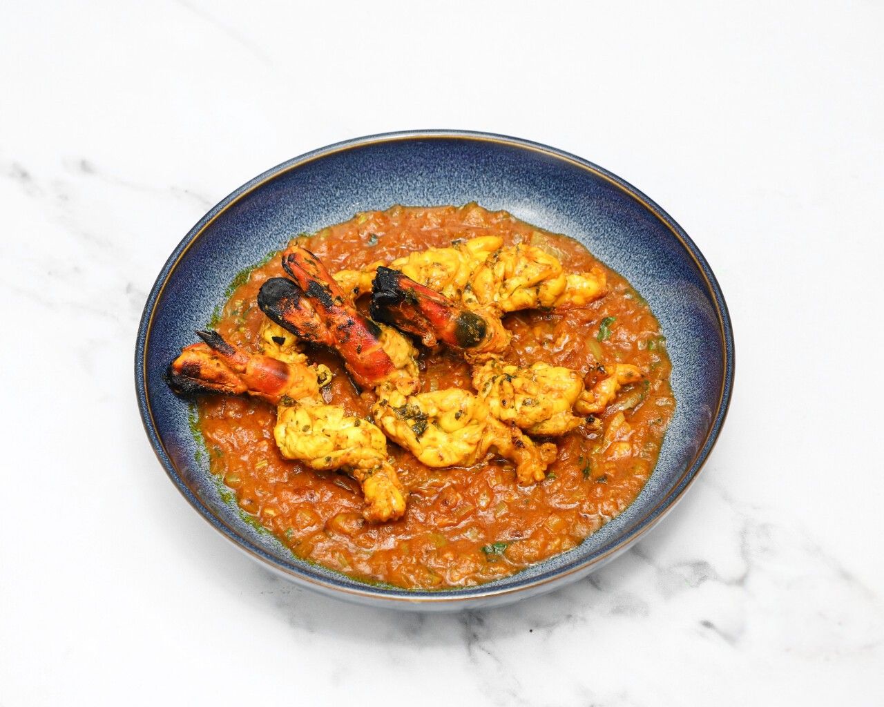 Tandoori king prawns with charred tails served in a blue bowl with a rich reddish-brown sauce.