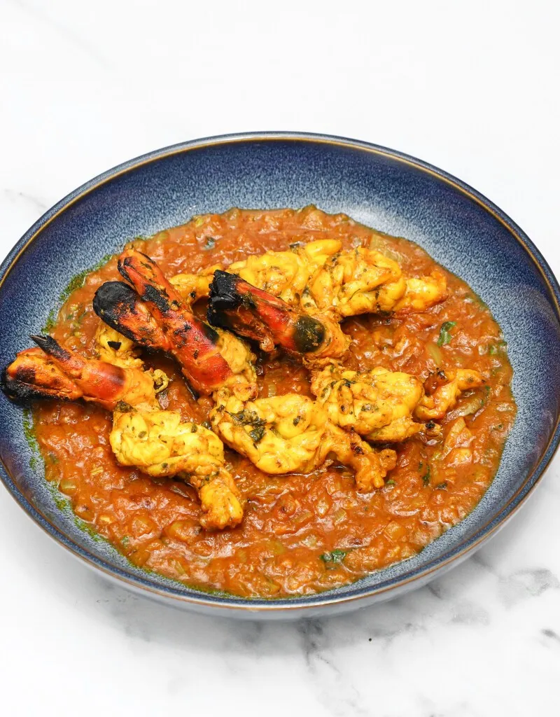 Tandoori king prawns with charred tails served in a blue bowl with a rich reddish-brown sauce.
