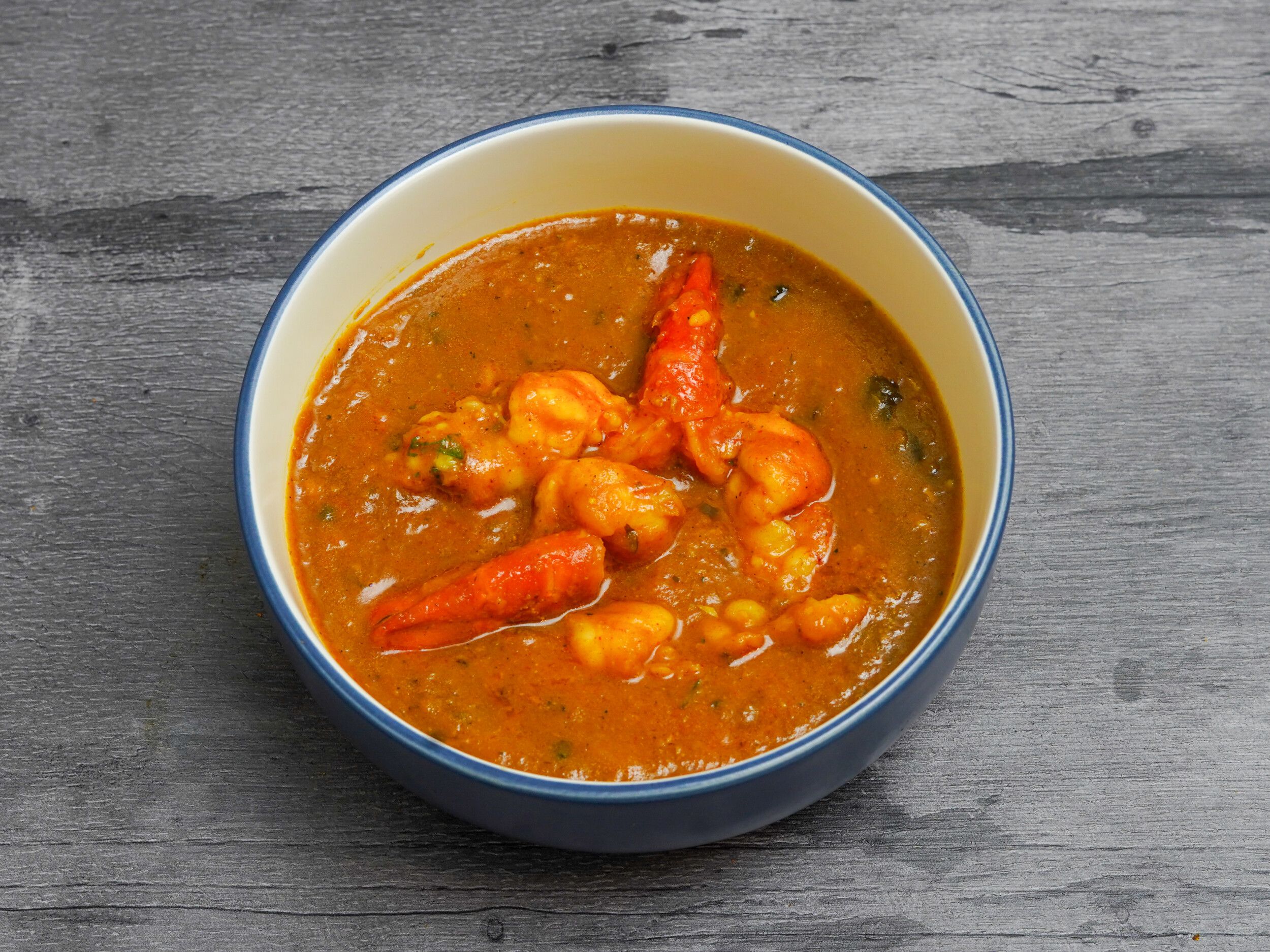 A blue and white bowl filled with an orange prawn curry, visible on a grey wooden surface