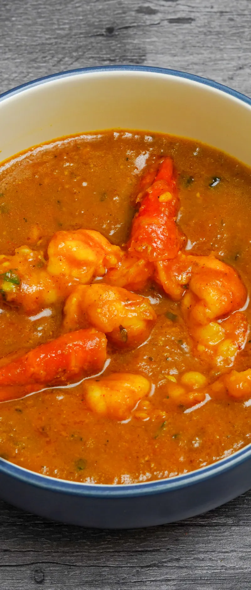 A blue and white bowl filled with an orange prawn curry, visible on a grey wooden surface