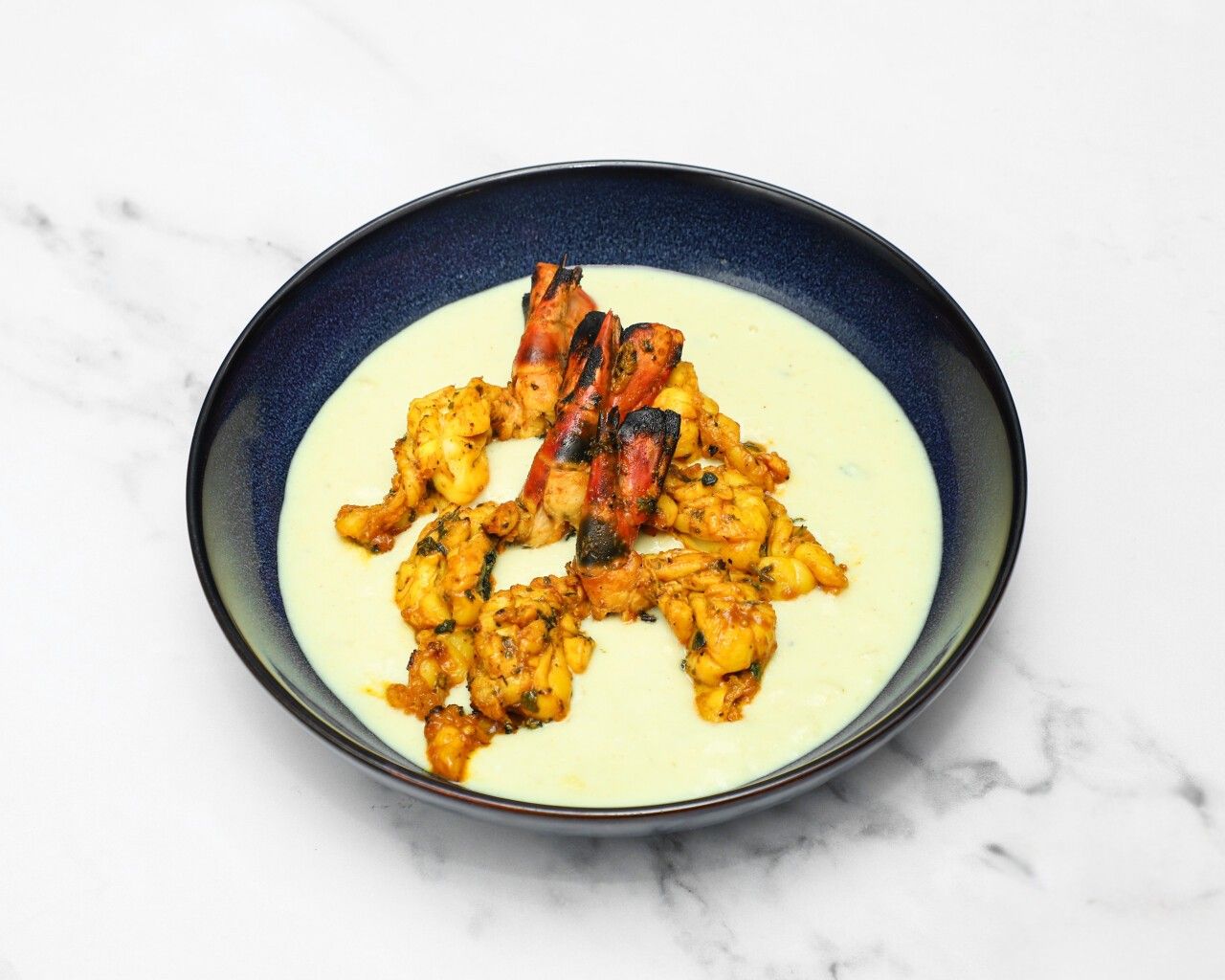 Grilled and spiced prawns and langoustines resting in a light yellow creamy sauce in a dark blue bowl.