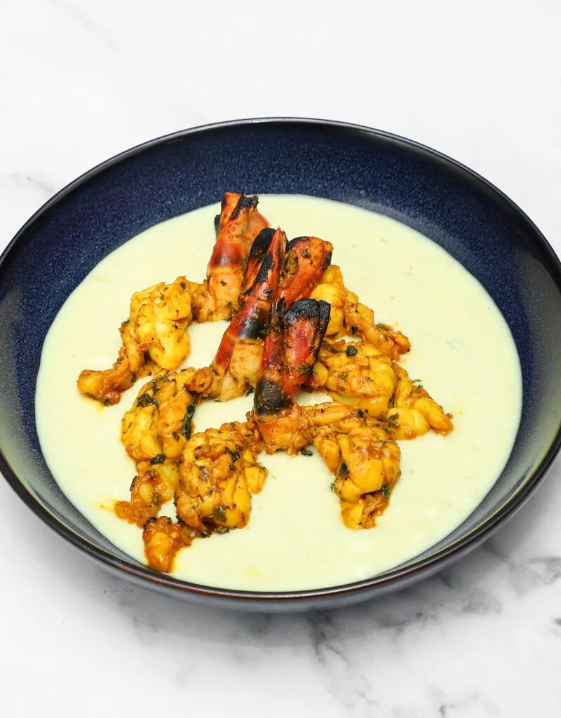 Grilled and spiced prawns and langoustines resting in a light yellow creamy sauce in a dark blue bowl.