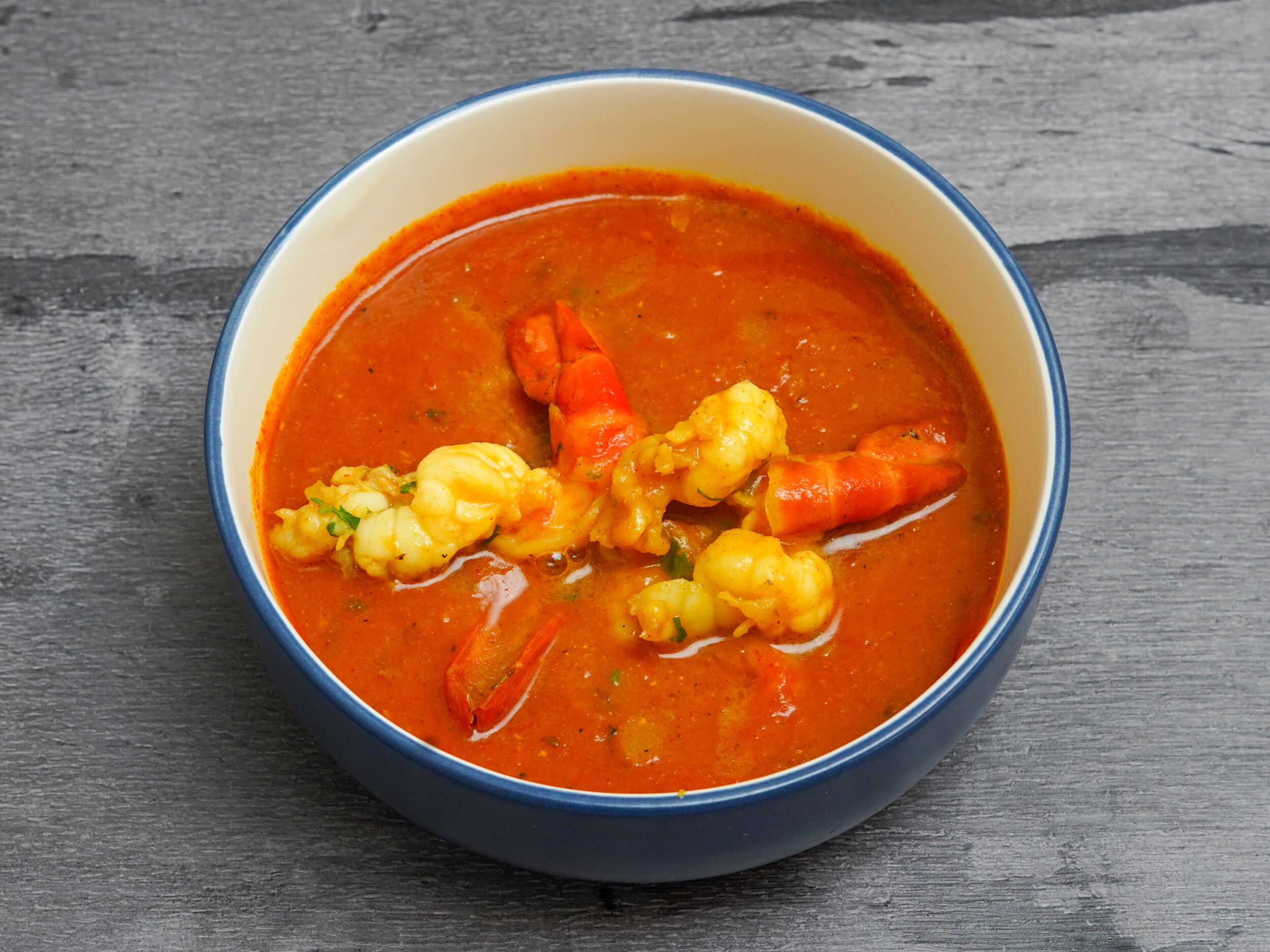 Bright orange prawn curry in a blue-rimmed white bowl on a gray wooden surface.