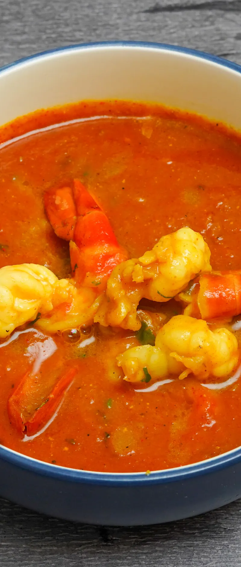 Bright orange prawn curry in a blue-rimmed white bowl on a gray wooden surface.