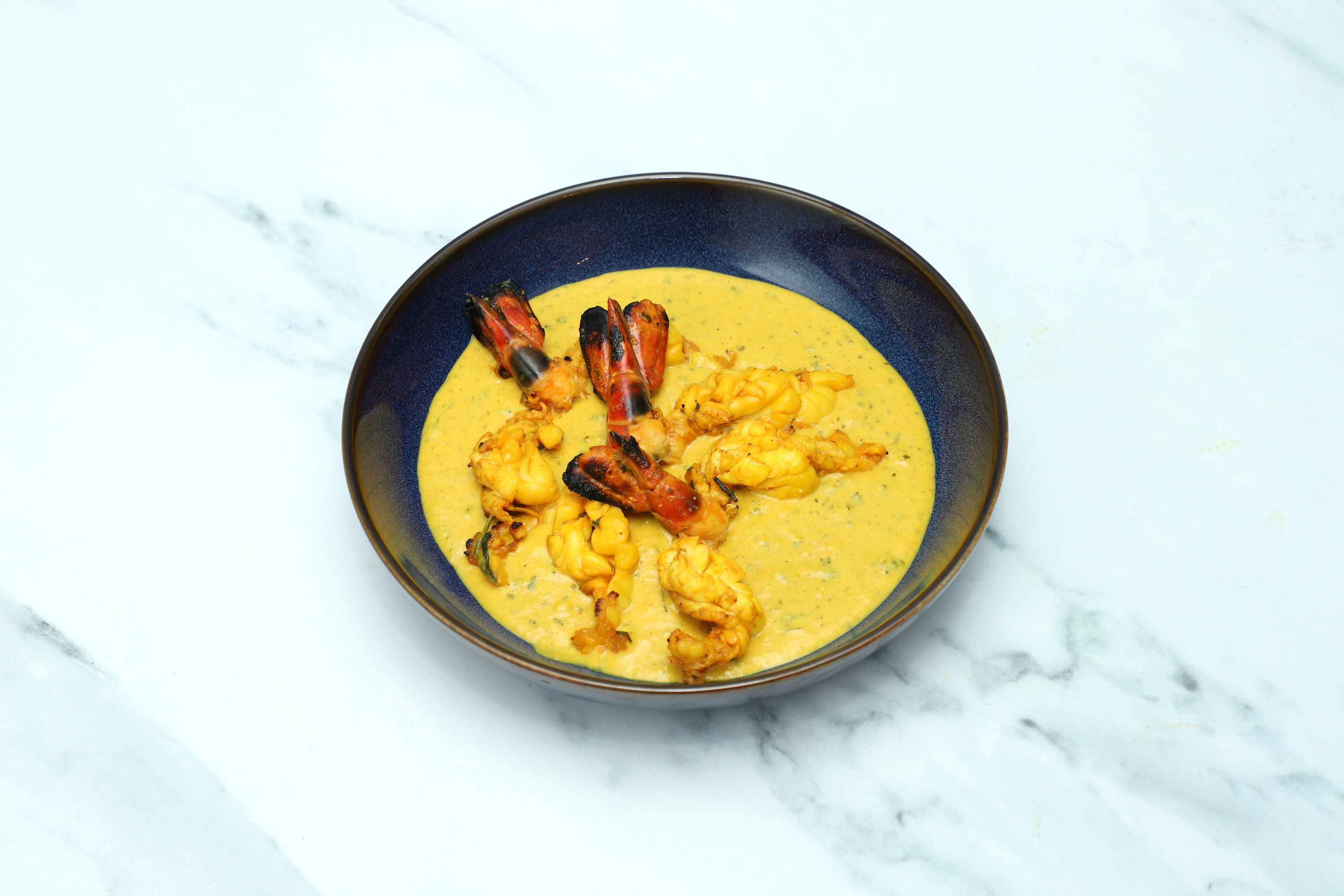 Grilled prawns with tails in a generous serving of yellow curried sauce in a dark blue bowl.