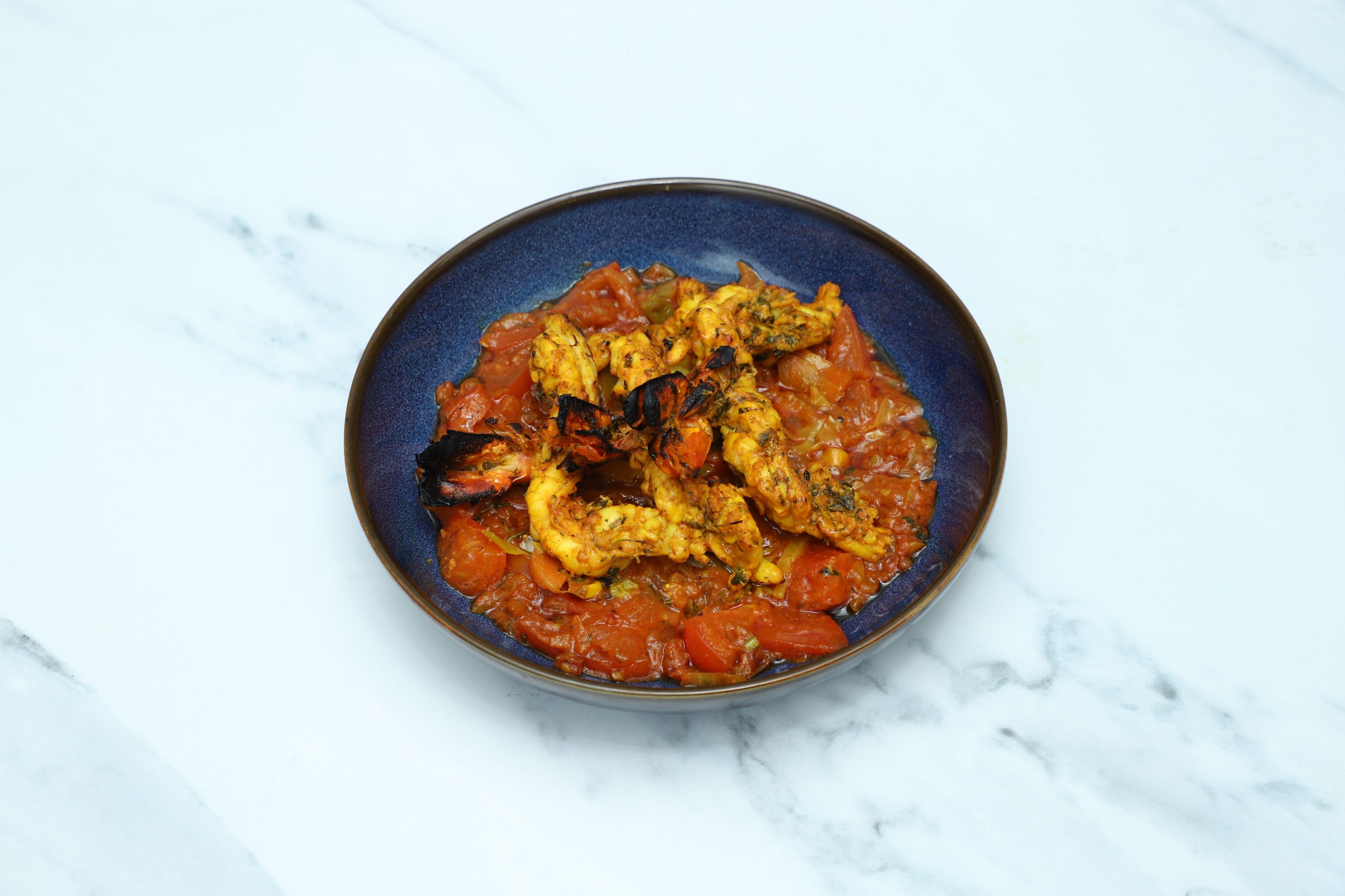 Grilled tandoori prawns are served atop a rich tomato and pepper masala in a vibrant blue bowl.