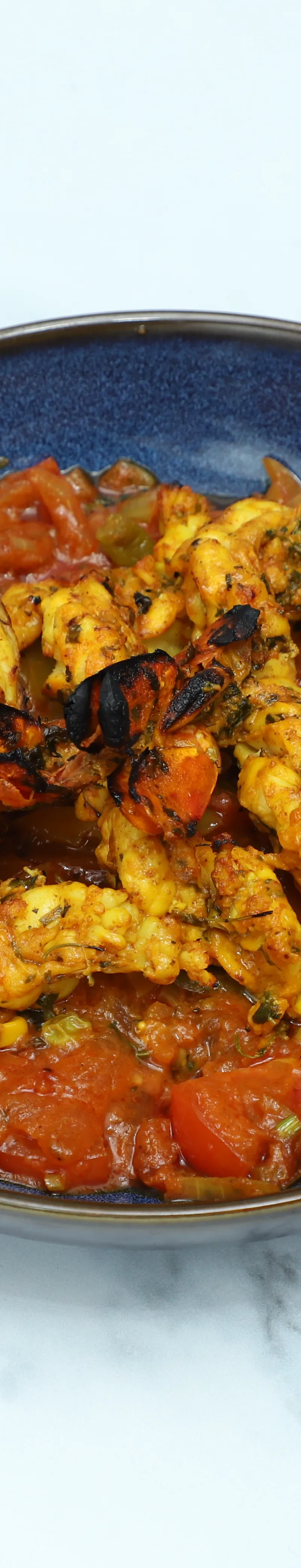 Grilled tandoori prawns are served atop a rich tomato and pepper masala in a vibrant blue bowl.