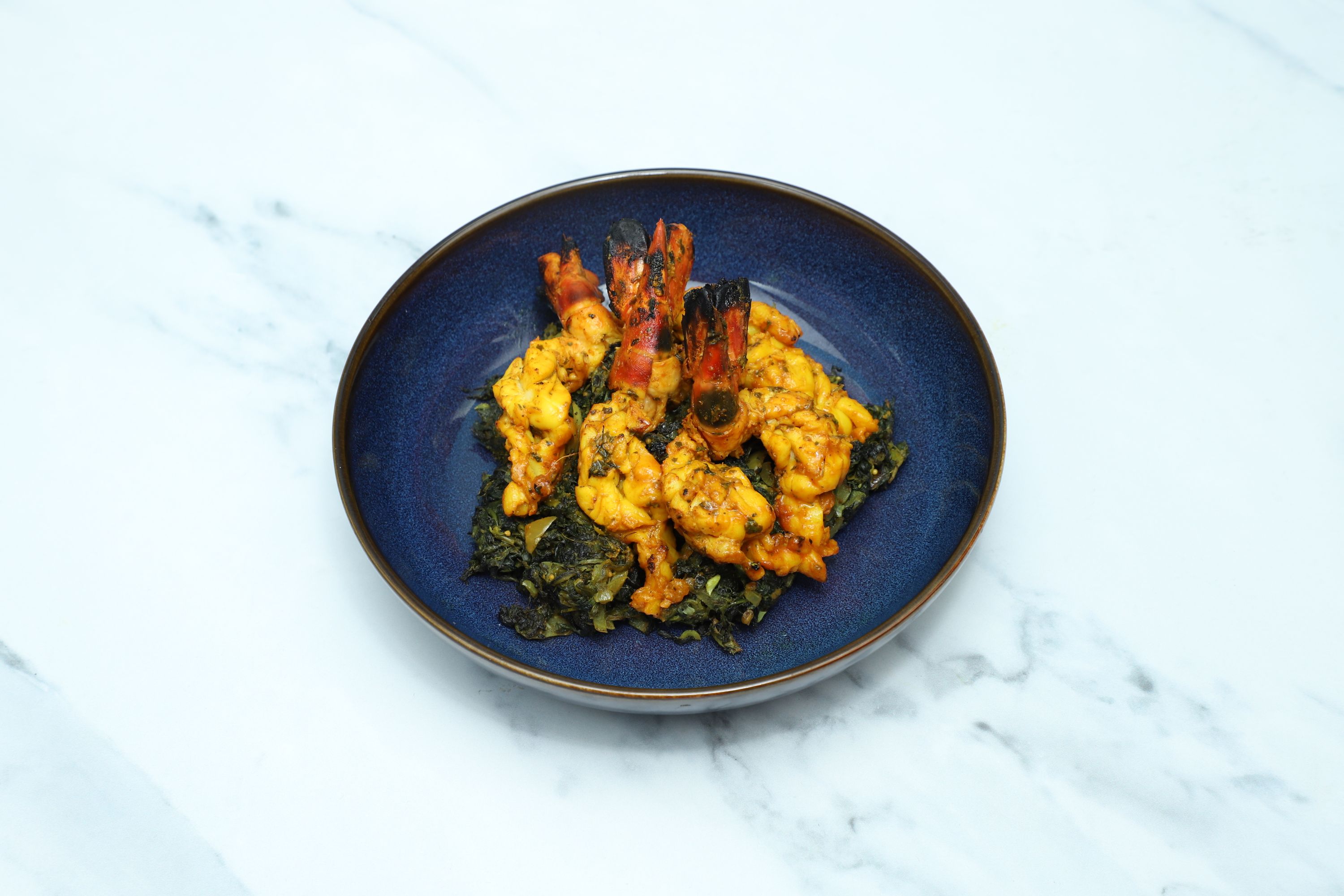 Grilled orange prawns and pale yellow pieces of chicken or fish serve on a bed of dark green Indian curried greens in a dark blue bowl.