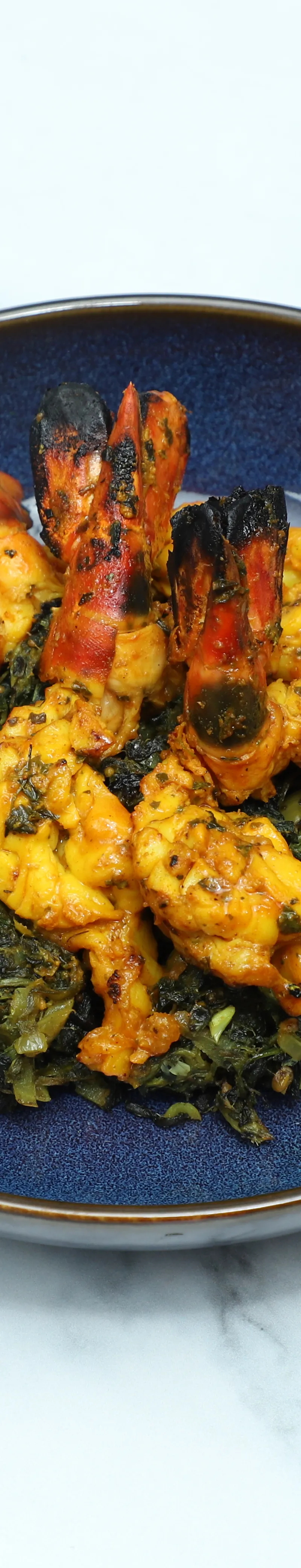 Grilled orange prawns and pale yellow pieces of chicken or fish serve on a bed of dark green Indian curried greens in a dark blue bowl.