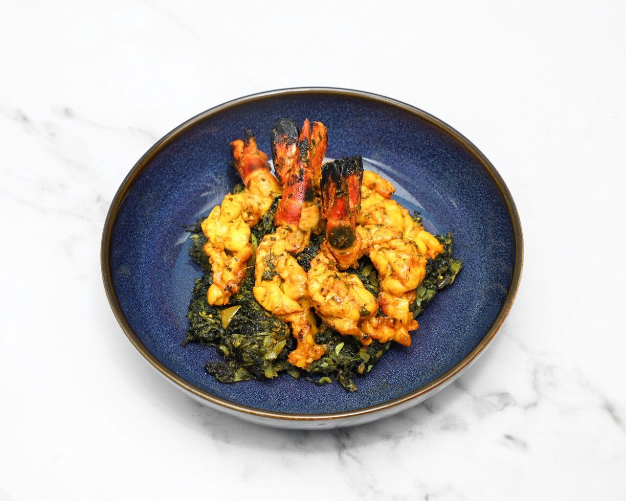 Grilled prawns with visible tails arranged on a bed of green saag in a deep blue bowl.