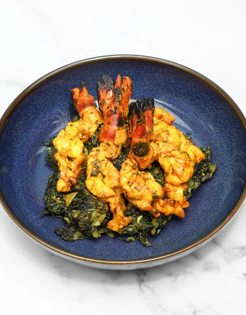 Grilled prawns with visible tails arranged on a bed of green saag in a deep blue bowl.