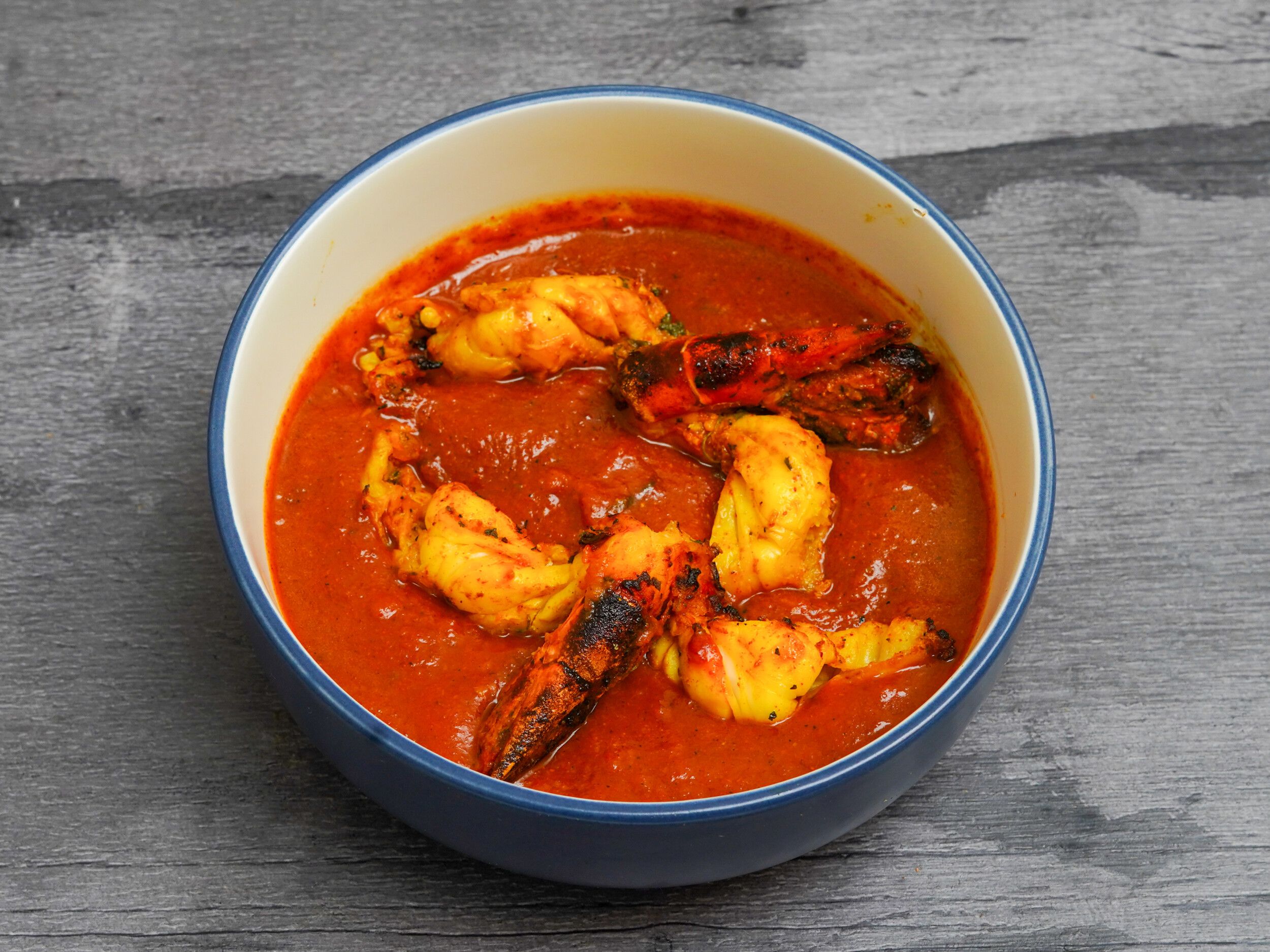 Several large king prawn butterfly pieces submerged in a vibrant orange-red curry sauce in a blue-rimmed bowl.