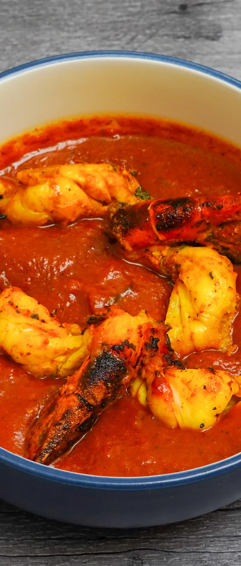 Several large king prawn butterfly pieces submerged in a vibrant orange-red curry sauce in a blue-rimmed bowl.