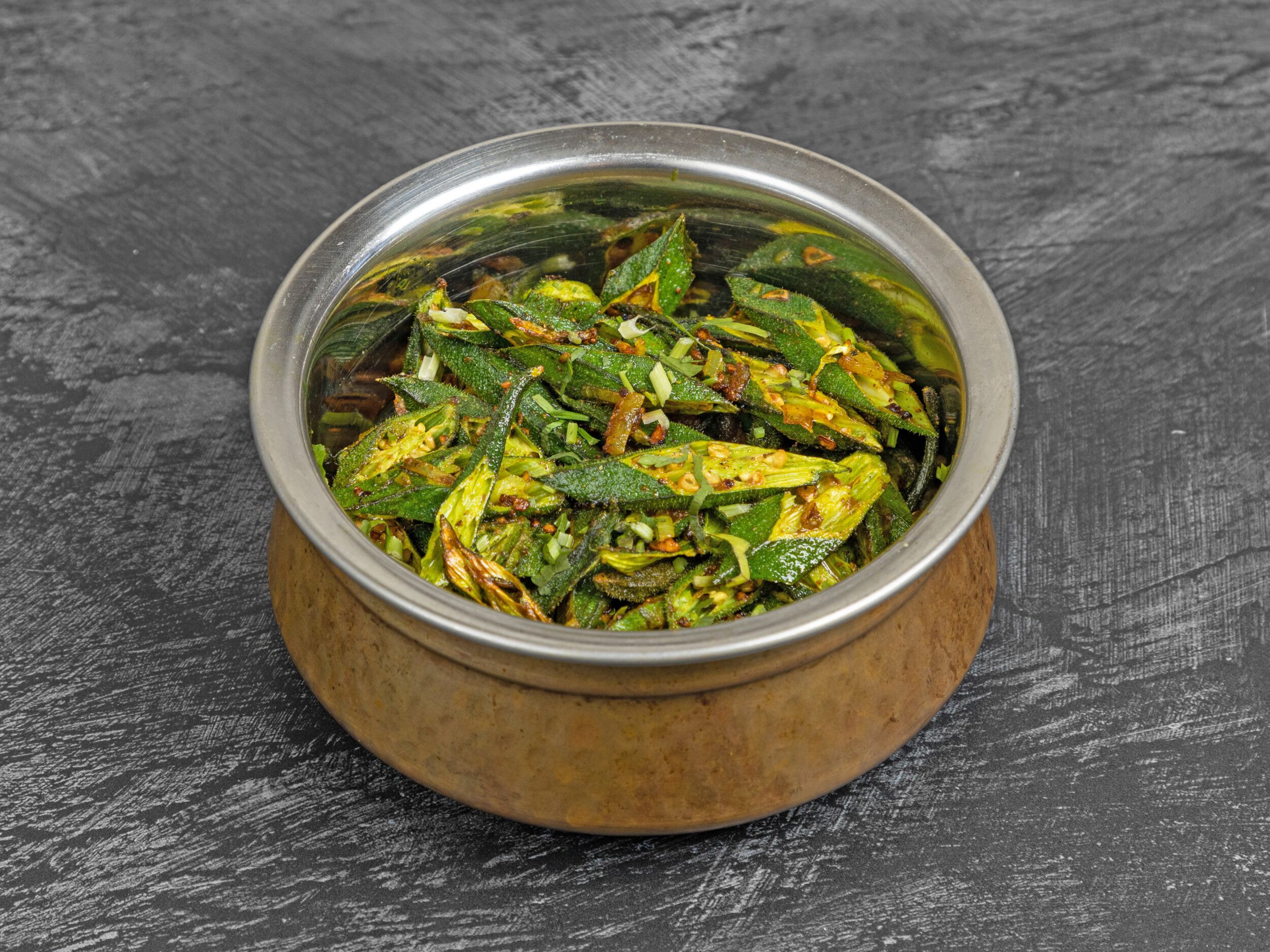 A copper karahi bowl filled with green bhindi fry (okra) on a dark textured surface.