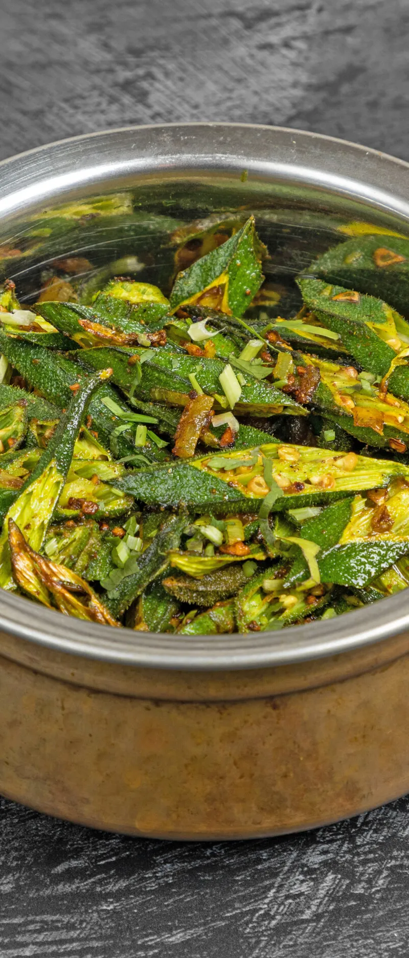A copper karahi bowl filled with green bhindi fry (okra) on a dark textured surface.