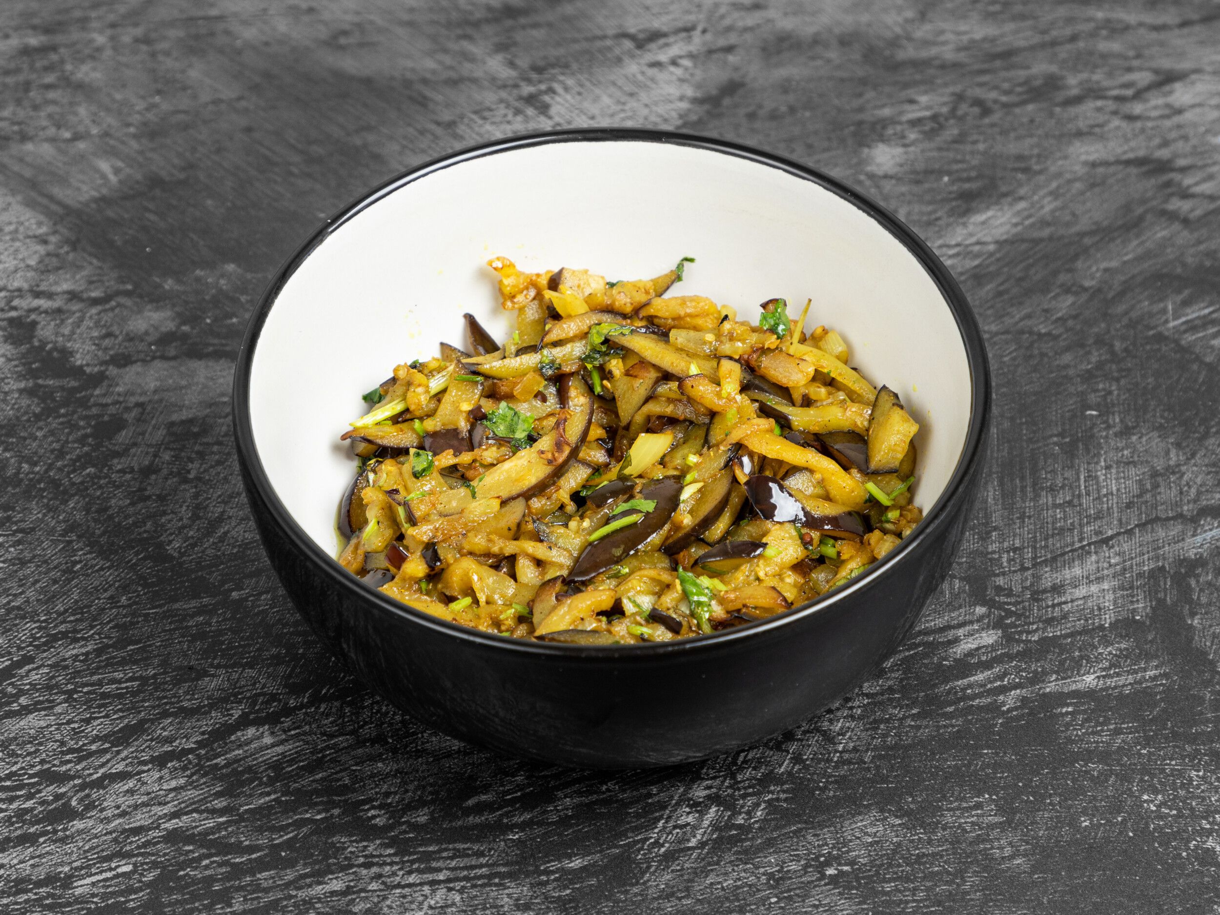 A black and white bowl filled with stir-fried aubergine slices and green herbs, on a dark textured surface.