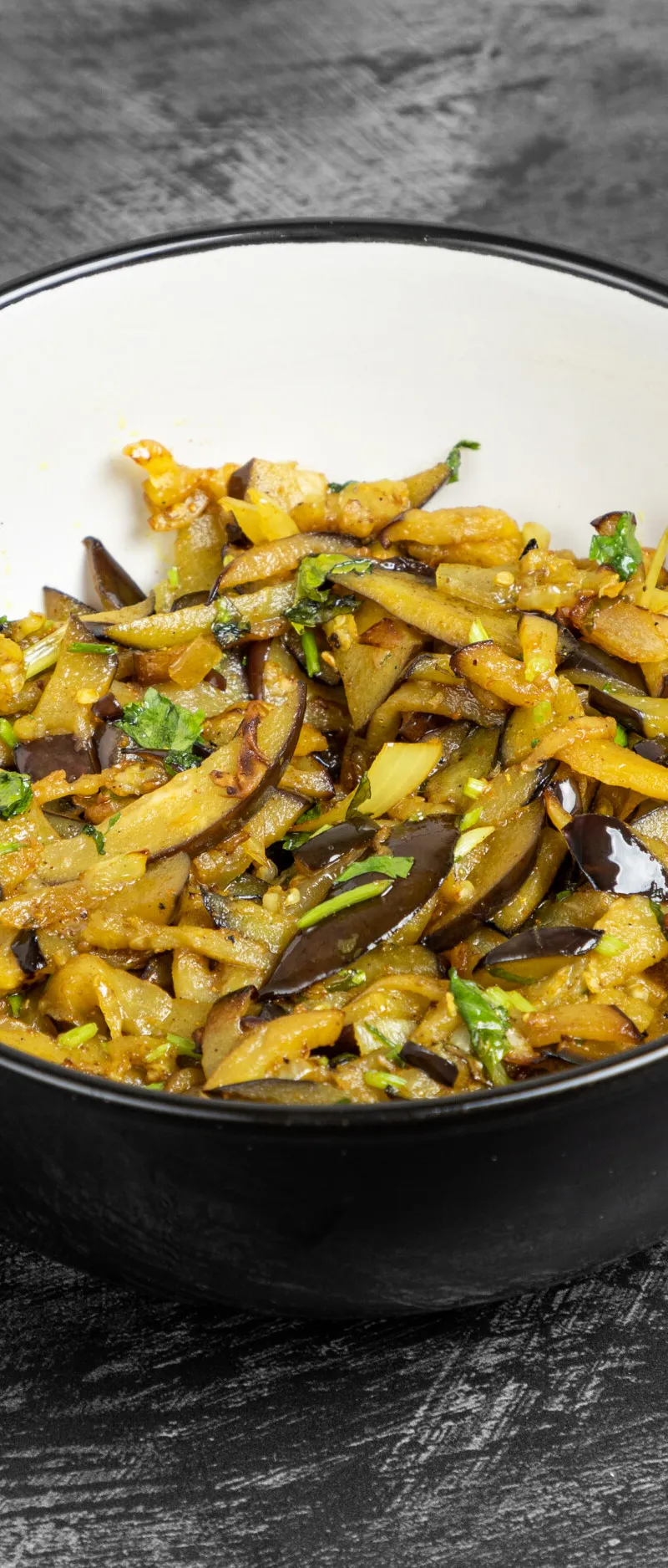 A black and white bowl filled with stir-fried aubergine slices and green herbs, on a dark textured surface.