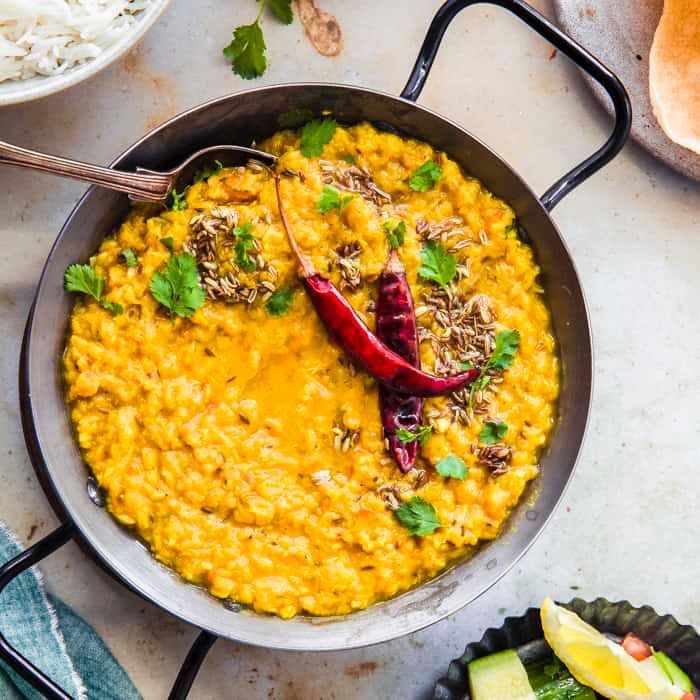 A large round serving pot filled with bright yellow daal, garnished with cumin seeds, dried red chillies, and fresh coriander.