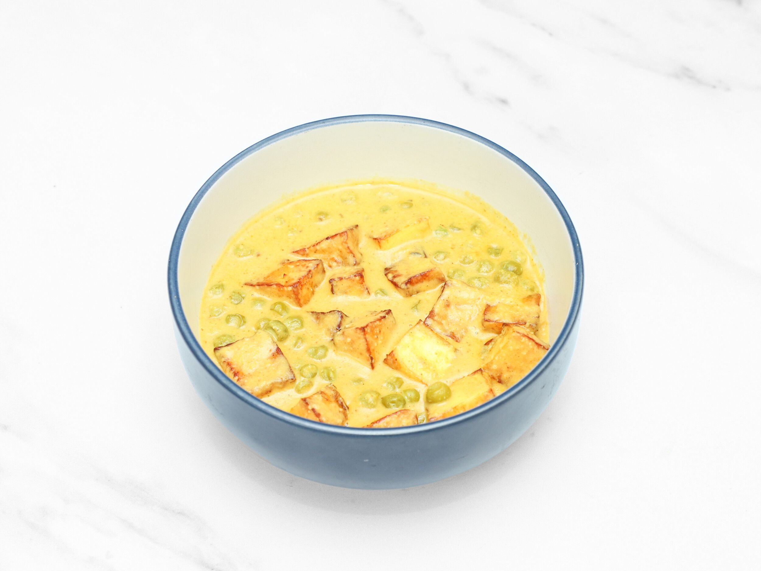 Mutter paneer, consisting of golden fried paneer cubes and green peas in a rich yellow sauce, in a blue bowl on a white table