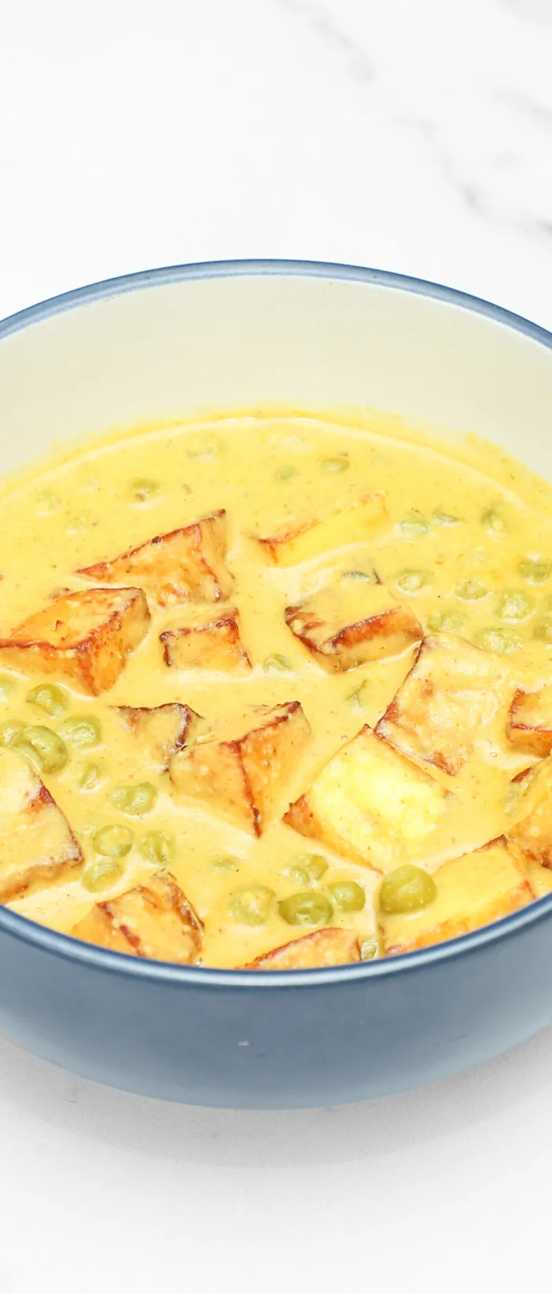 Mutter paneer, consisting of golden fried paneer cubes and green peas in a rich yellow sauce, in a blue bowl on a white table