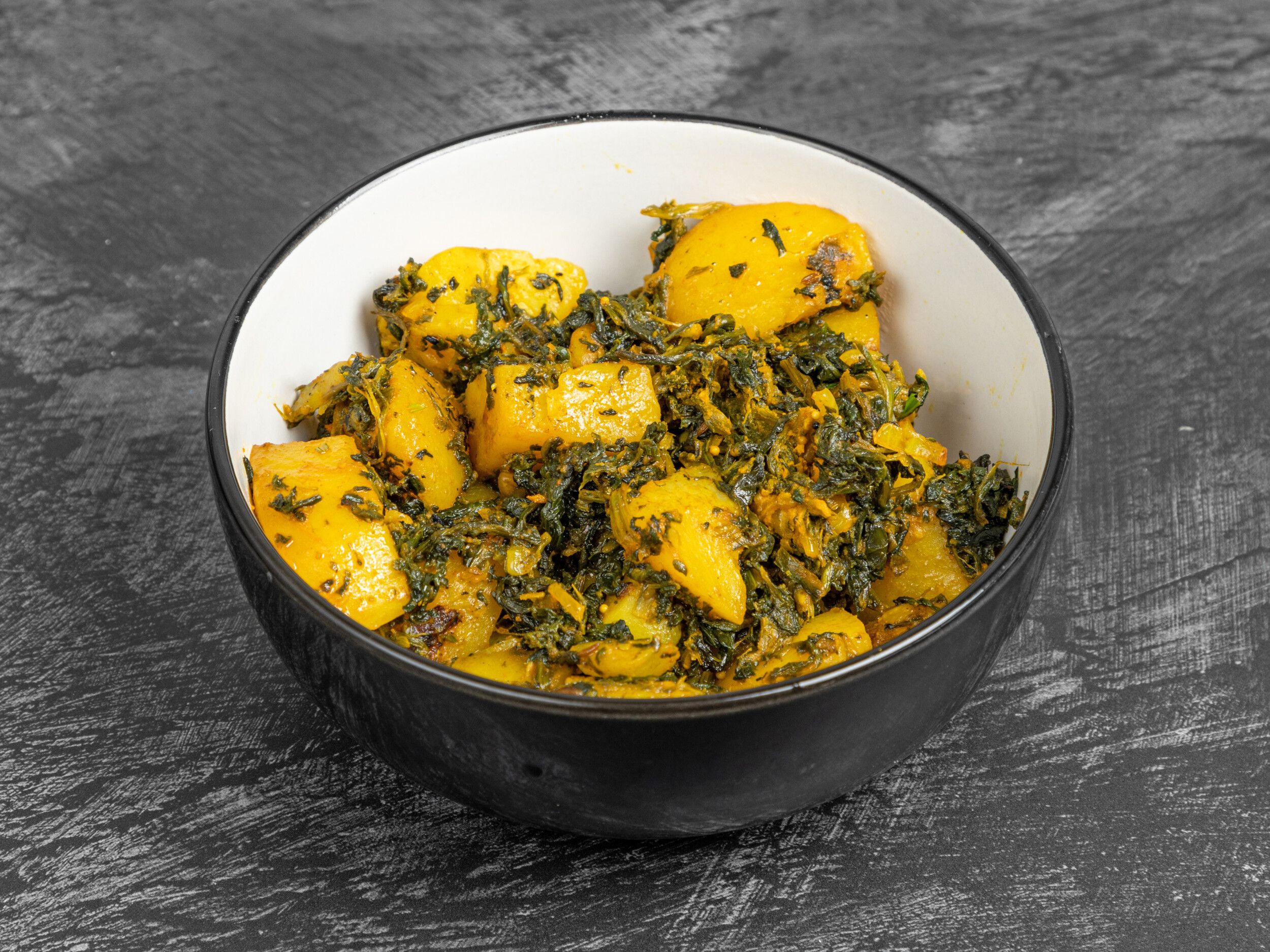 A black bowl filled with Aloo Saag, featuring vibrant yellow potato chunks and green spinach in a rich curry sauce, on a dark textured background.