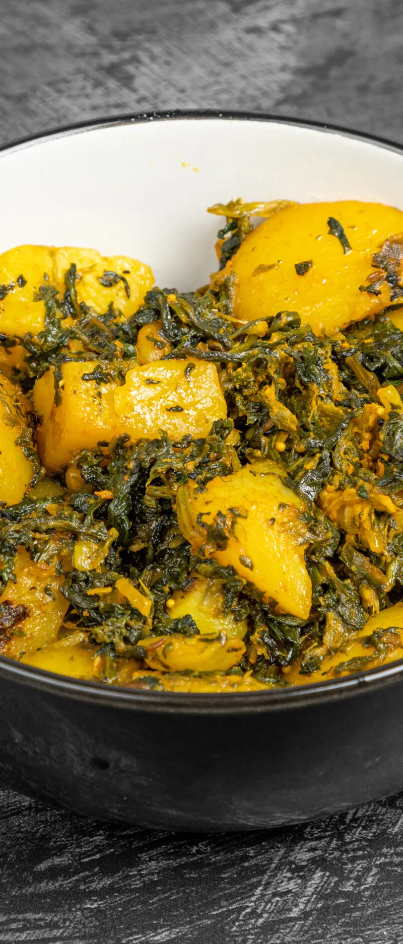A black bowl filled with Aloo Saag, featuring vibrant yellow potato chunks and green spinach in a rich curry sauce, on a dark textured background.