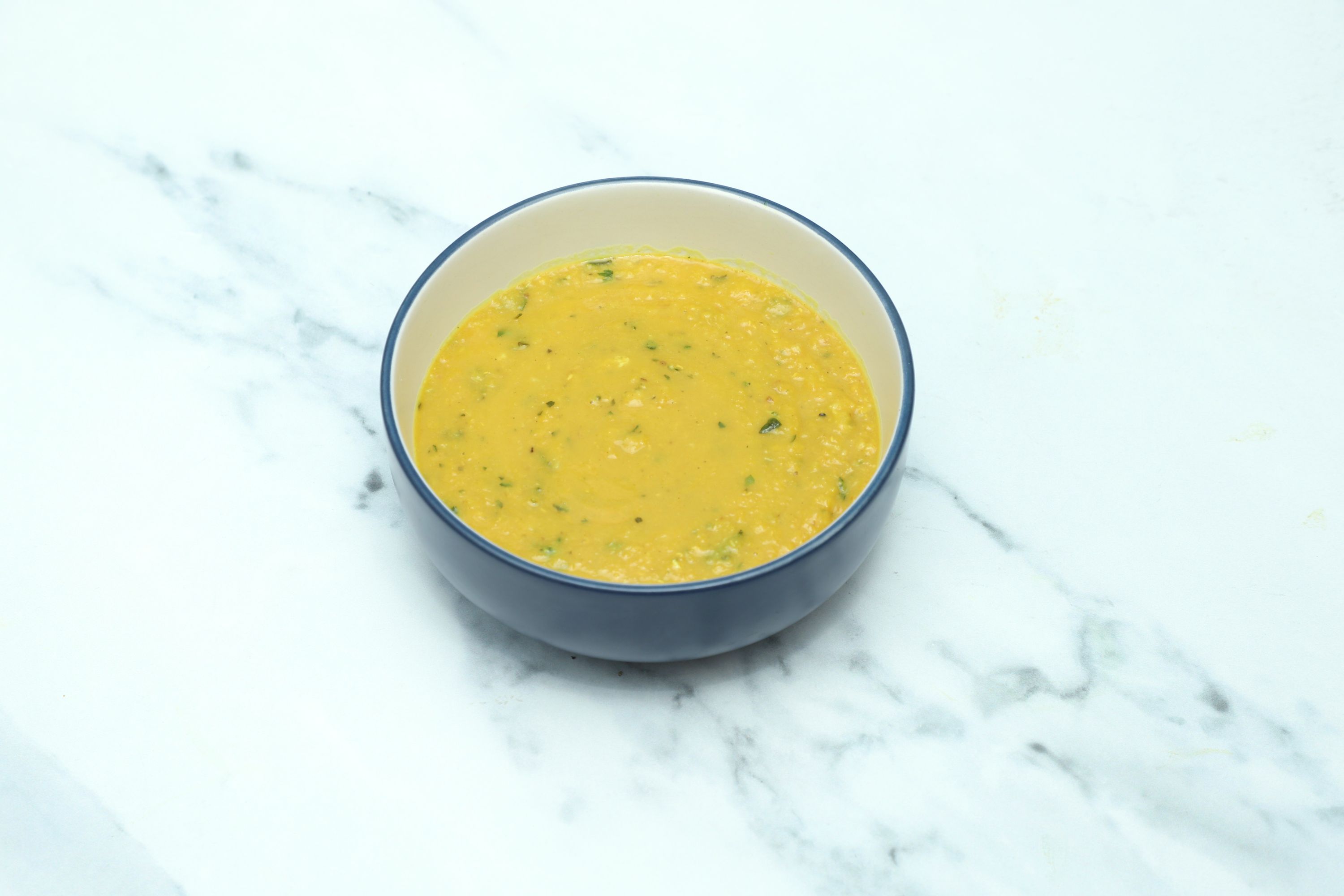 A round blue and white bowl filled with creamy yellow daal, garnished with green herbs, on a white marble surface.