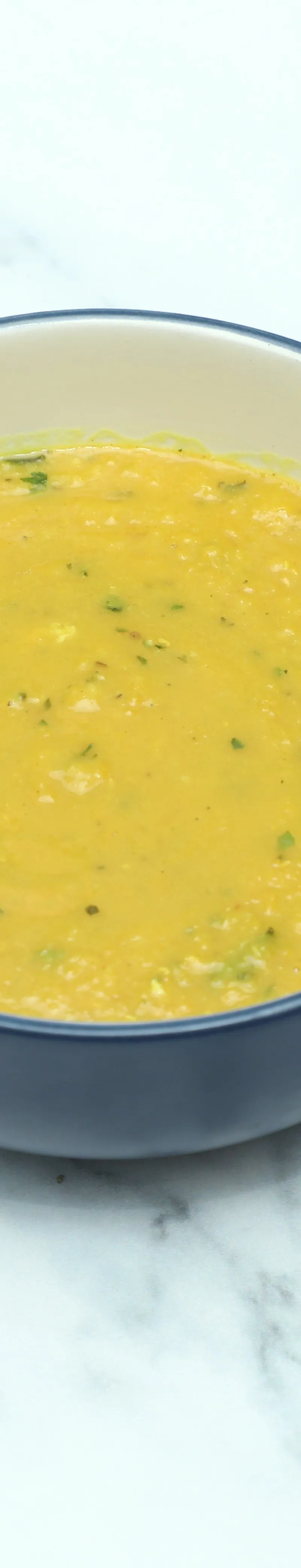 A round blue and white bowl filled with creamy yellow daal, garnished with green herbs, on a white marble surface.