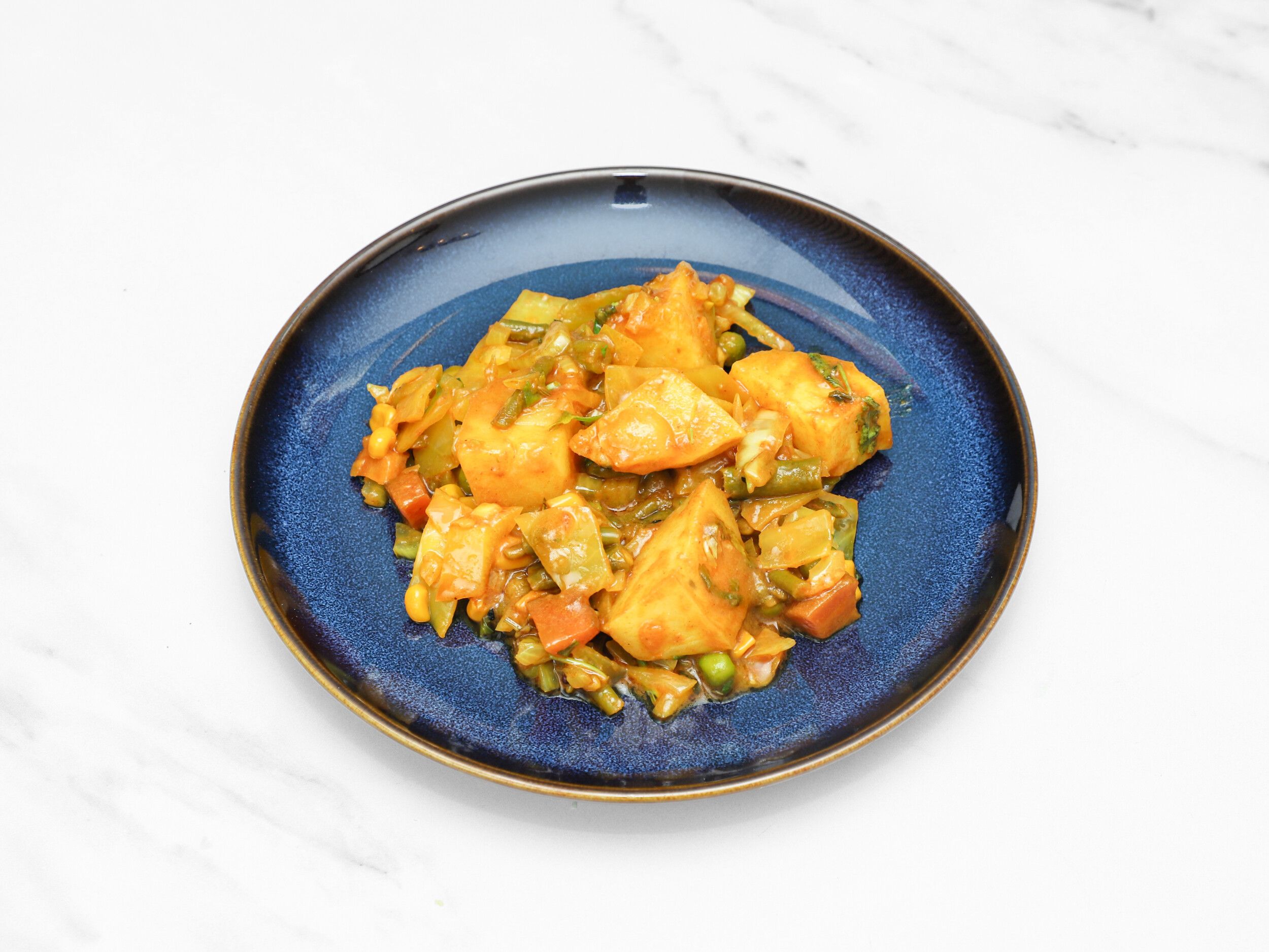 Mixed vegetable curry with potatoes, green beans, carrots, and peas on a navy blue plate.
