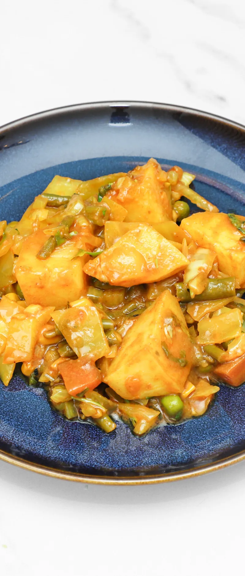 Mixed vegetable curry with potatoes, green beans, carrots, and peas on a navy blue plate.