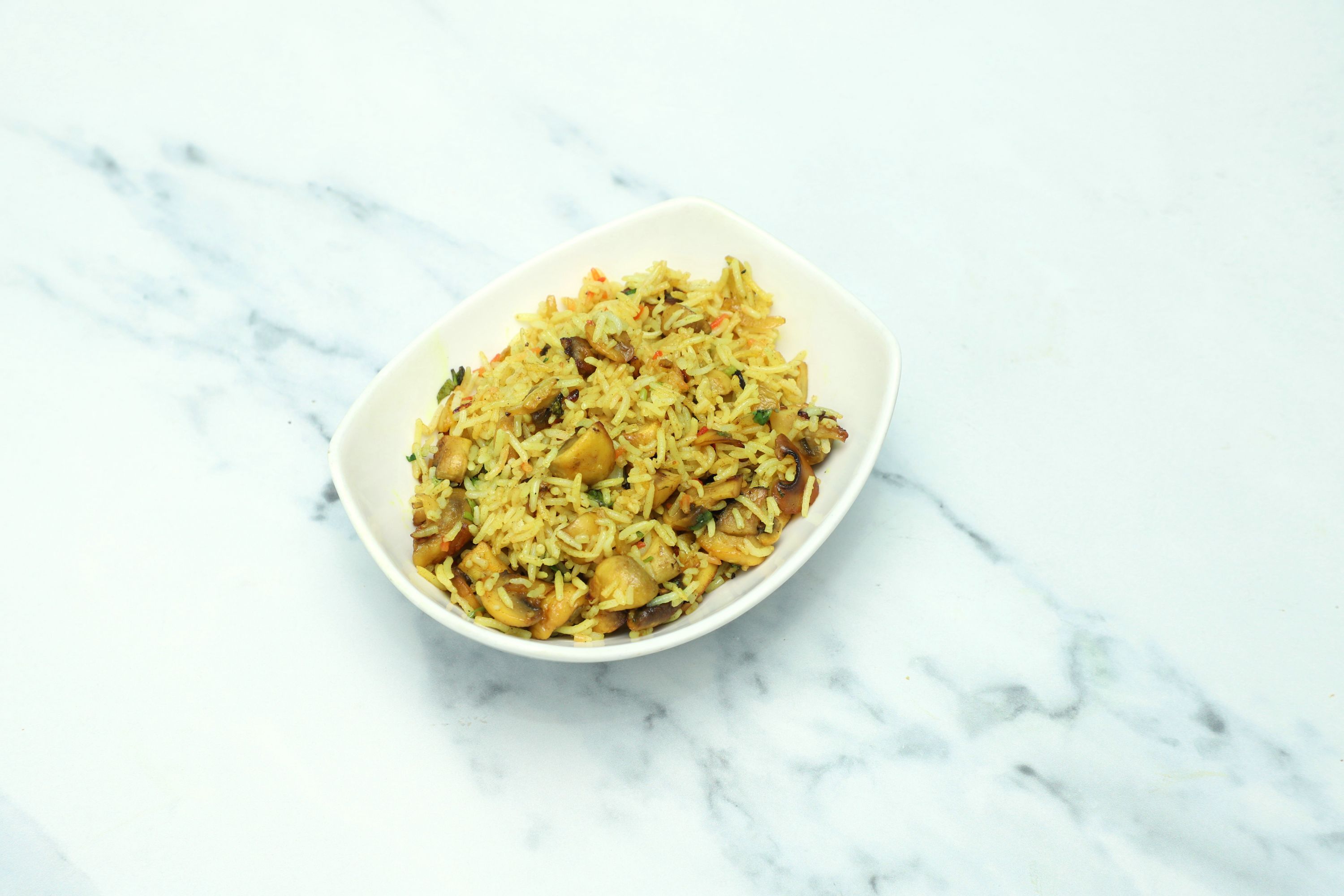 Mushroom pilau in a white, elongated bowl on a light marble surface. Rice is yellow with visible mushrooms.