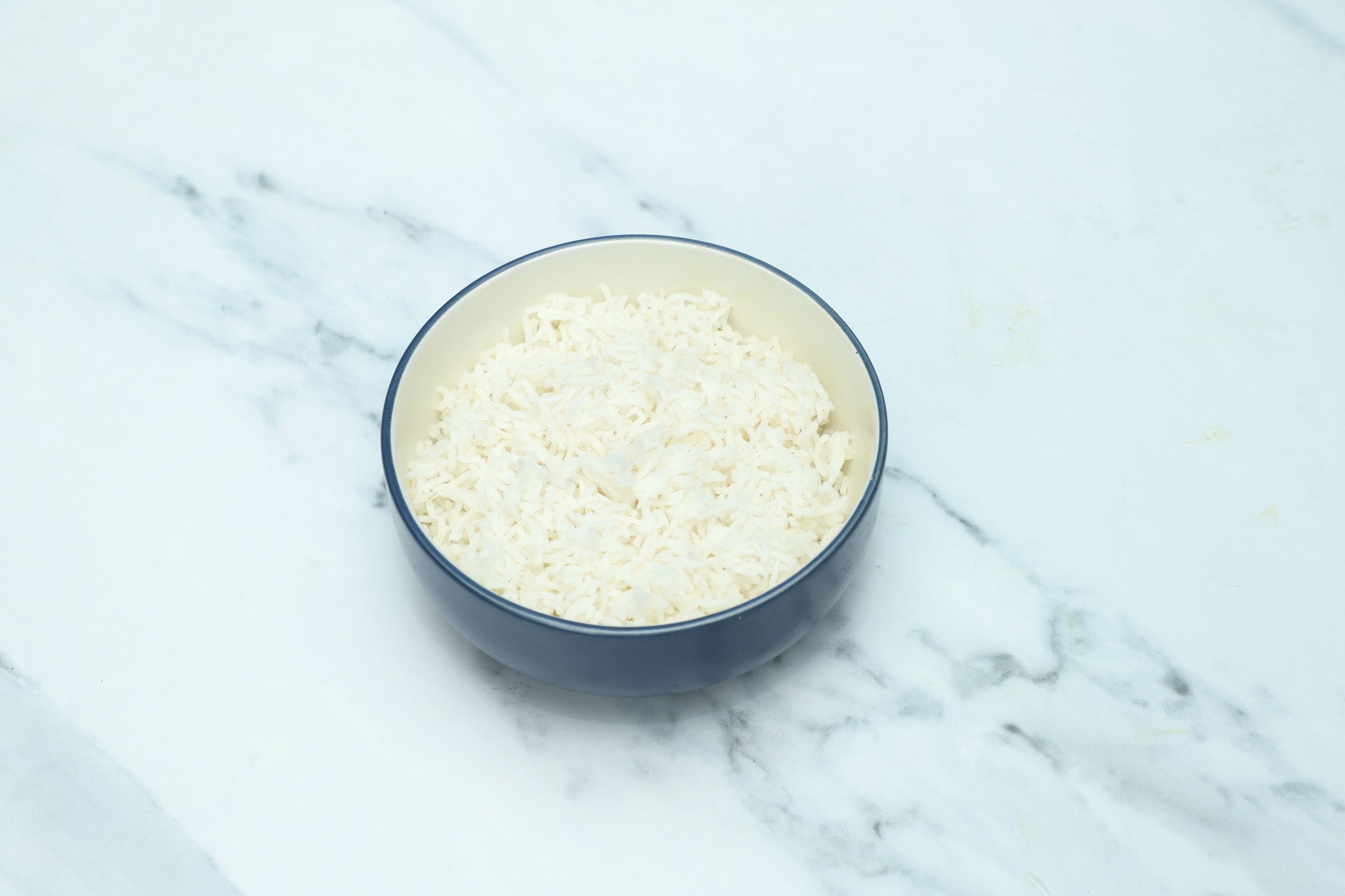 A round, dark blue and cream bowl filled with fluffy white basmati rice on a white marble surface.